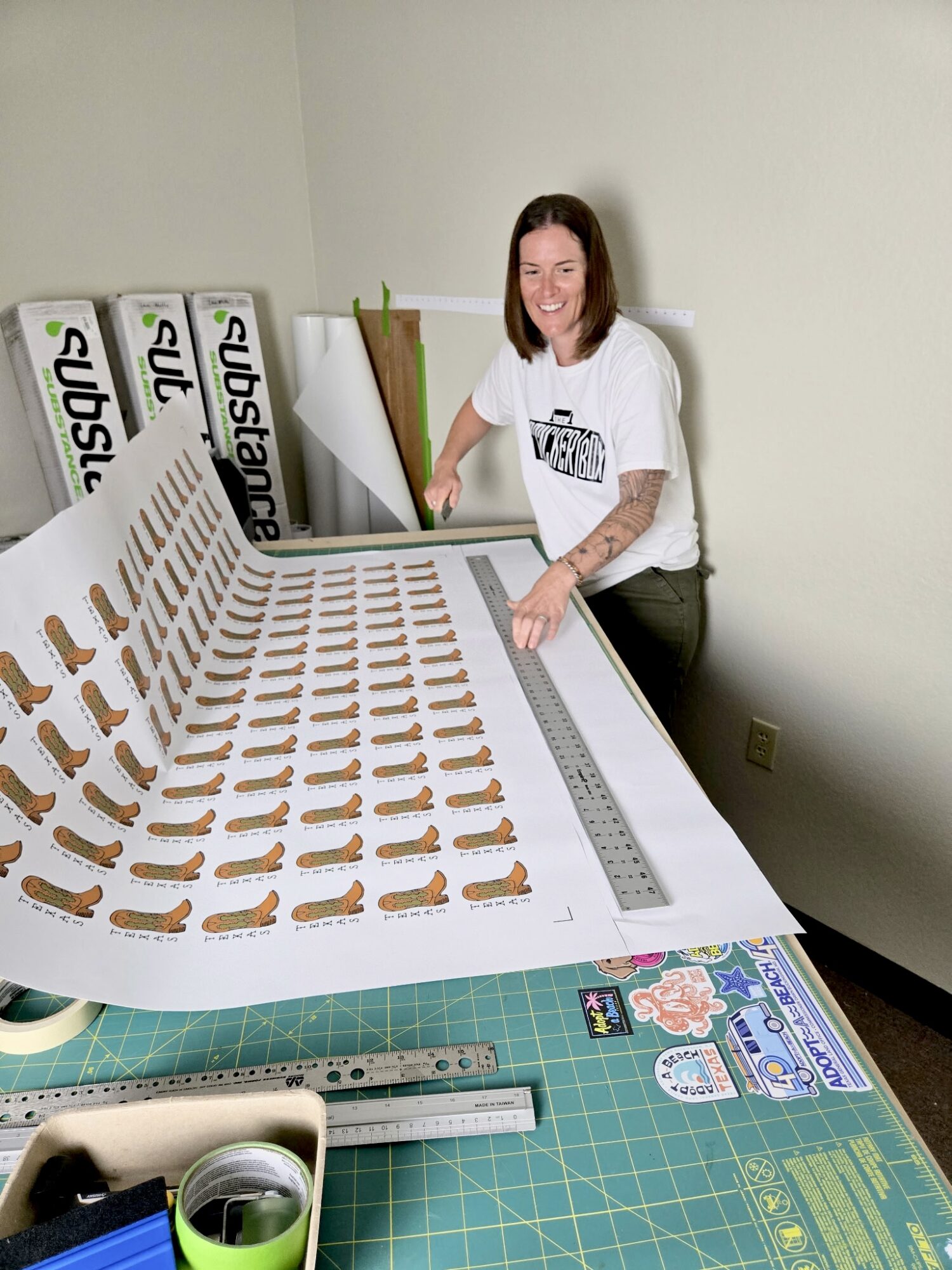 Woman standing at a table with numerous paper cutouts, measuring tools, and a cutting mat, smiling at the camera.
