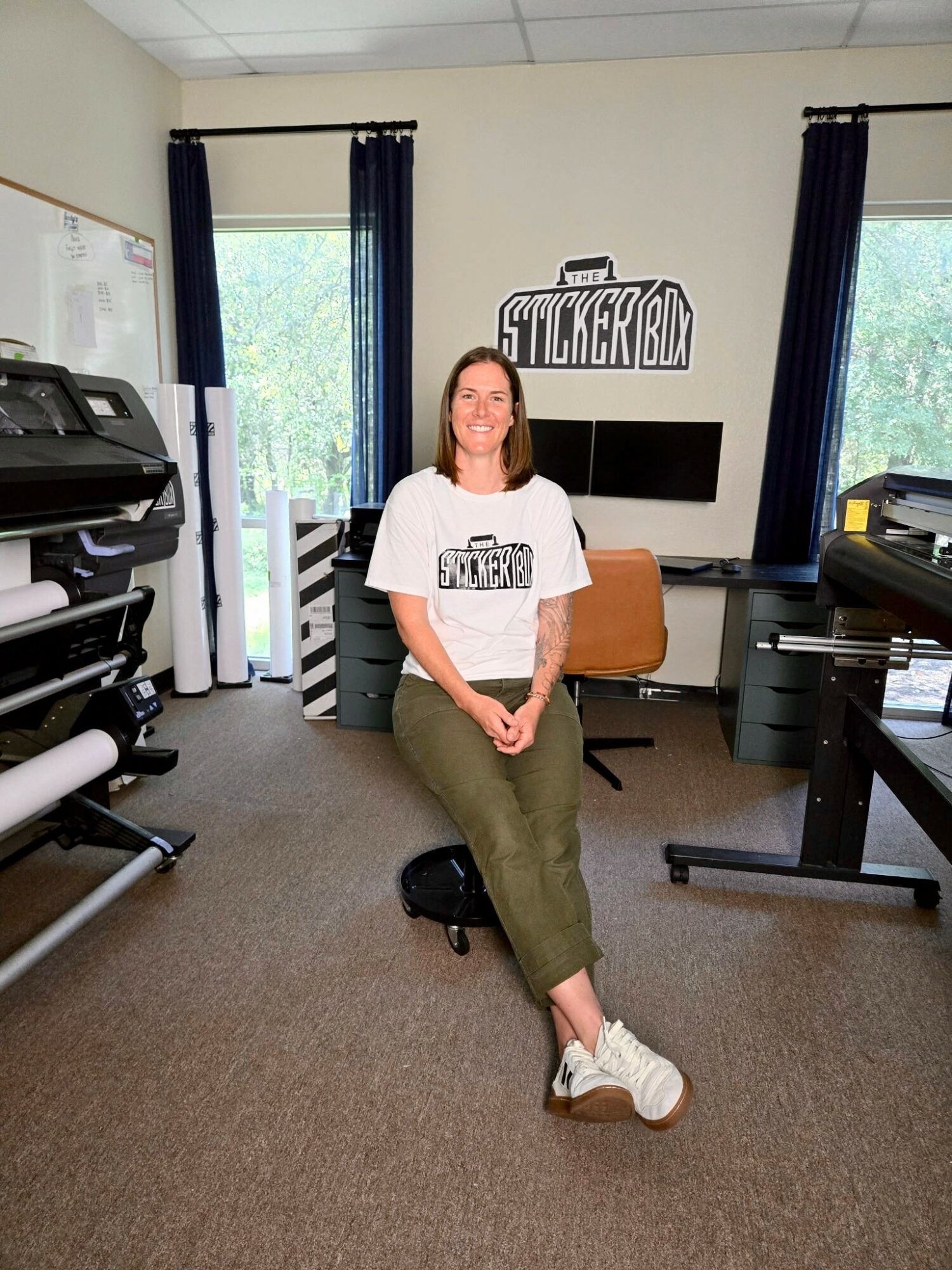 Woman sitting on a chair in an office with windows, black curtains, and a sticker box sign on the wall.