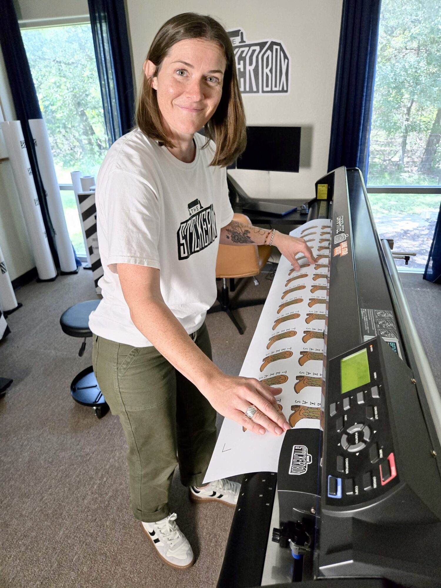 Woman with shoulder-length brown hair smiling at keyboard, wearing a white T-shirt and green pants, in a room with large windows.