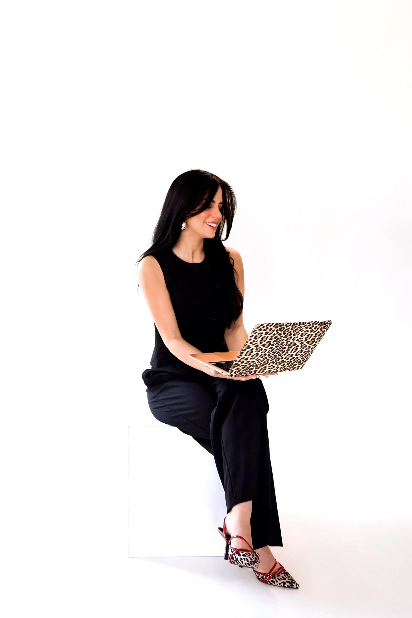 Woman sitting and smiling while using a laptop with a leopard print cover, wearing a black sleeveless top and pants, against a white background.