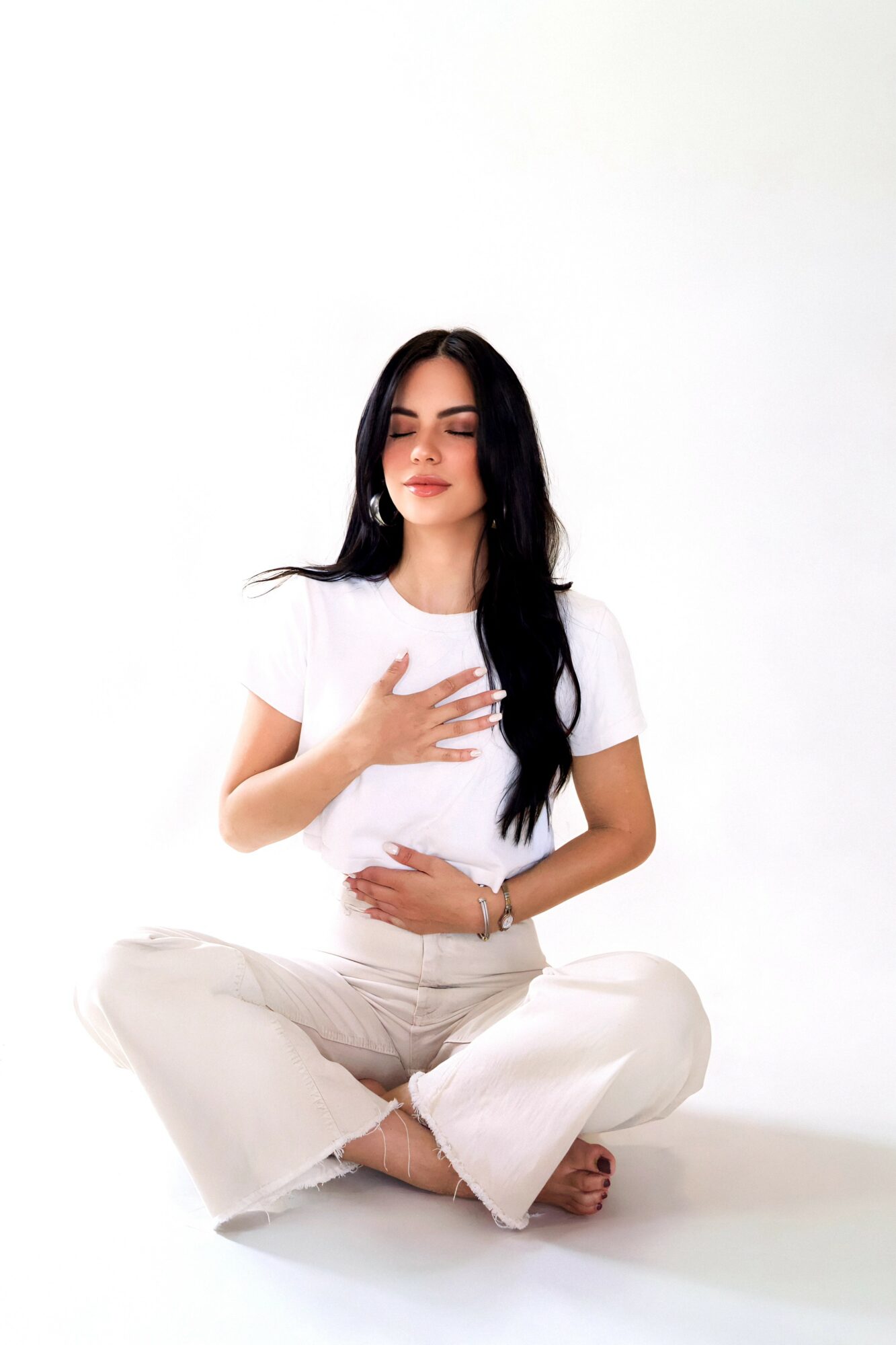 Woman sitting cross-legged with eyes closed, hand on chest, in a white outfit against a plain white background.