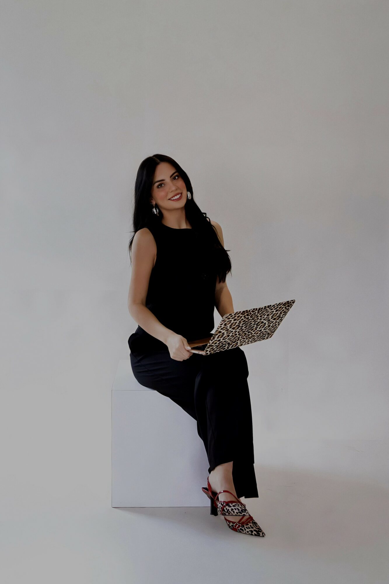 Woman sitting on a white block, holding a laptop, smiling, against a plain light background.