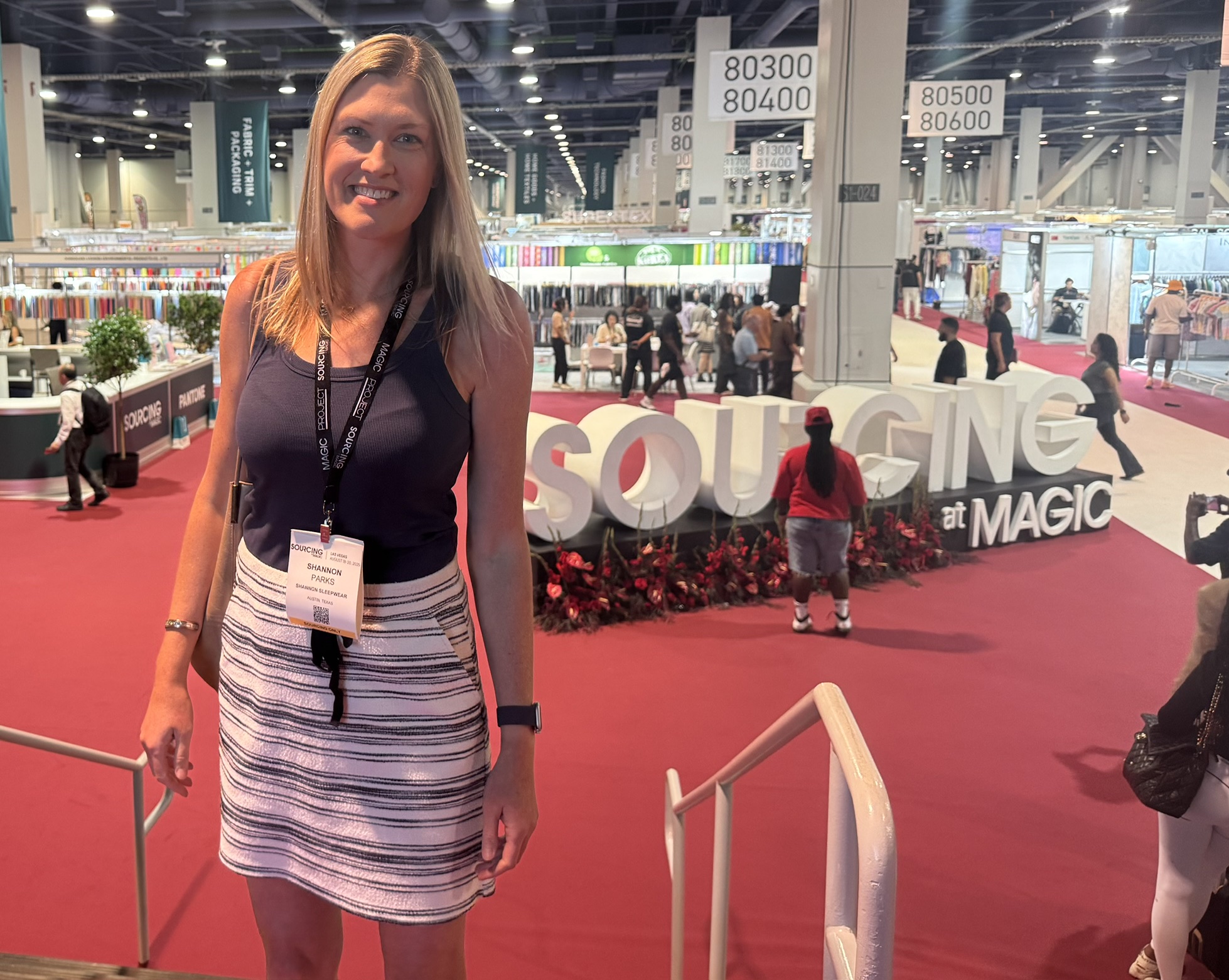 Woman with blonde hair wearing a black top and striped skirt, standing indoors at an event with booths and signs in the background.