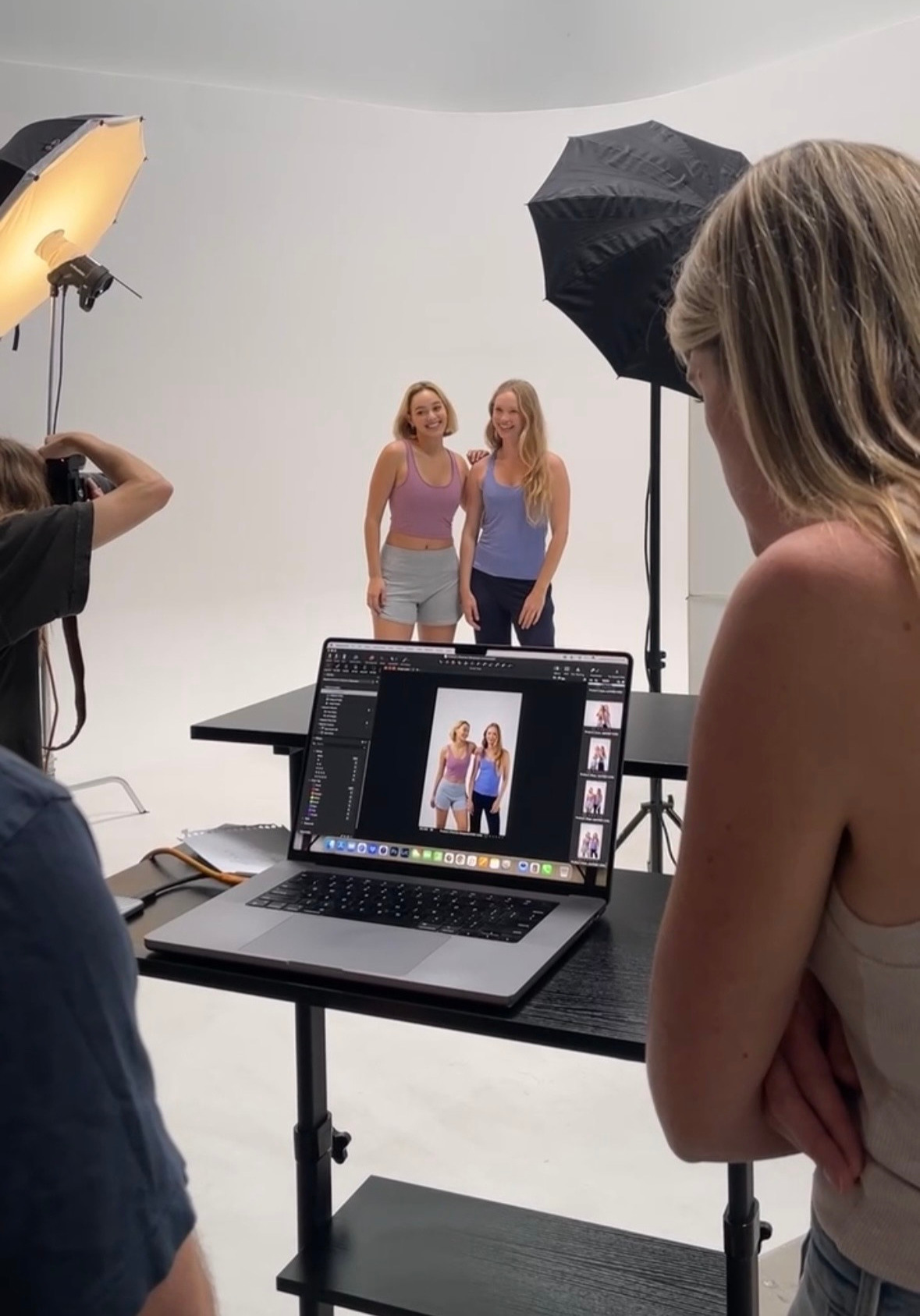 Two women stand in front of a white backdrop during a photo shoot, with a photographer and lighting equipment present.