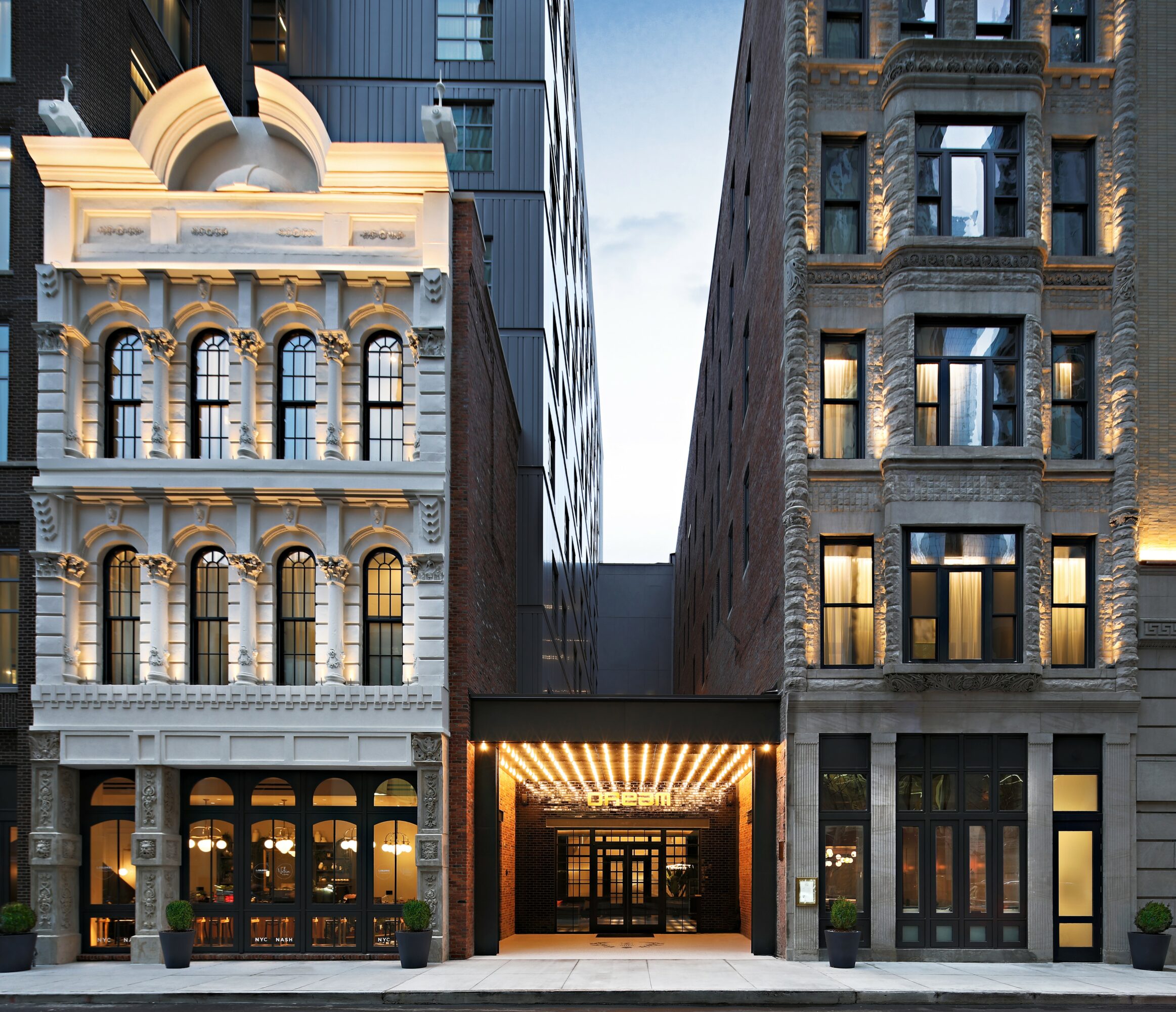 Two tall buildings with lit windows and an illuminated entrance between them, with a cloudy sky above.