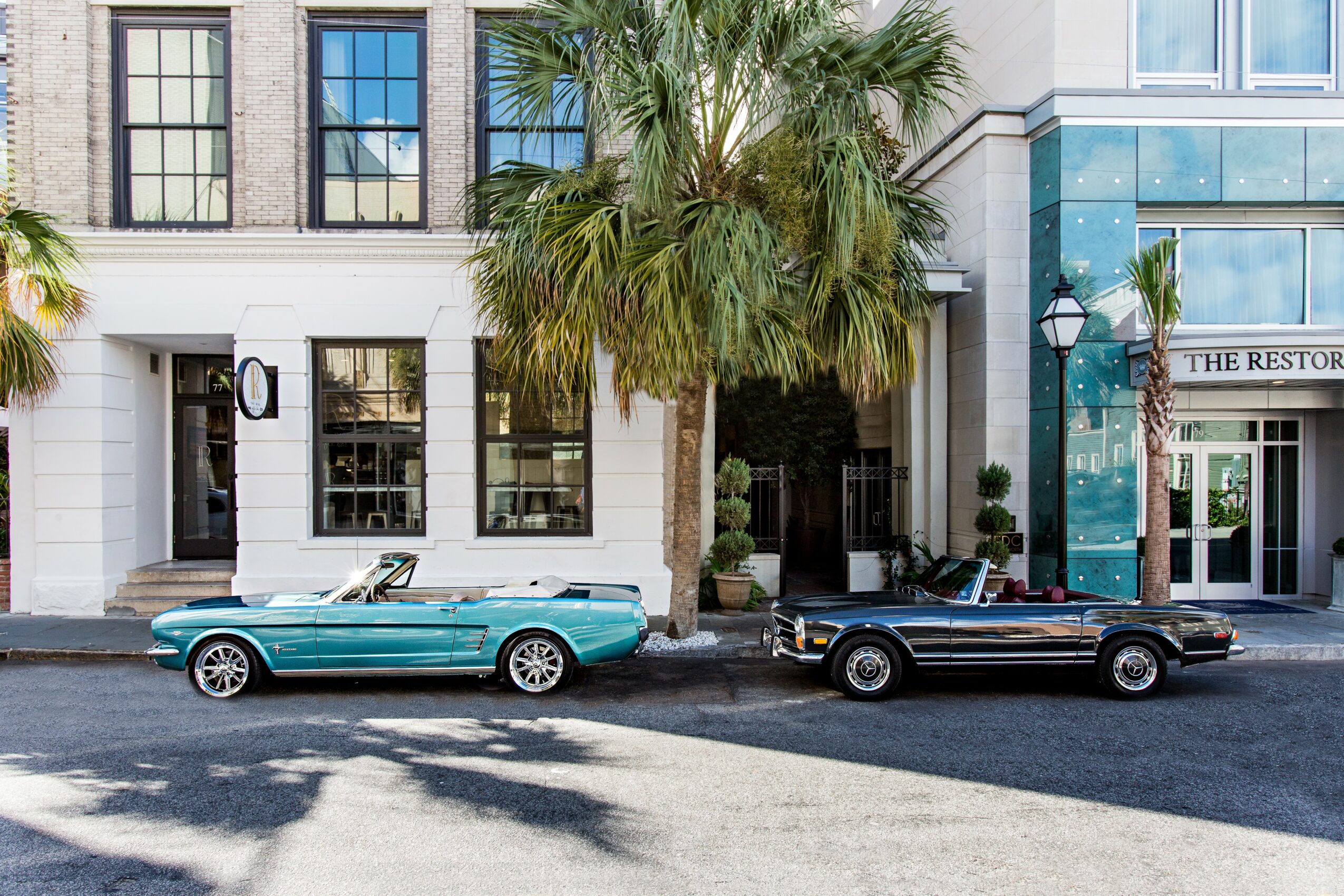 Two convertible cars parked on a street in front of a building with large windows and a palm tree.