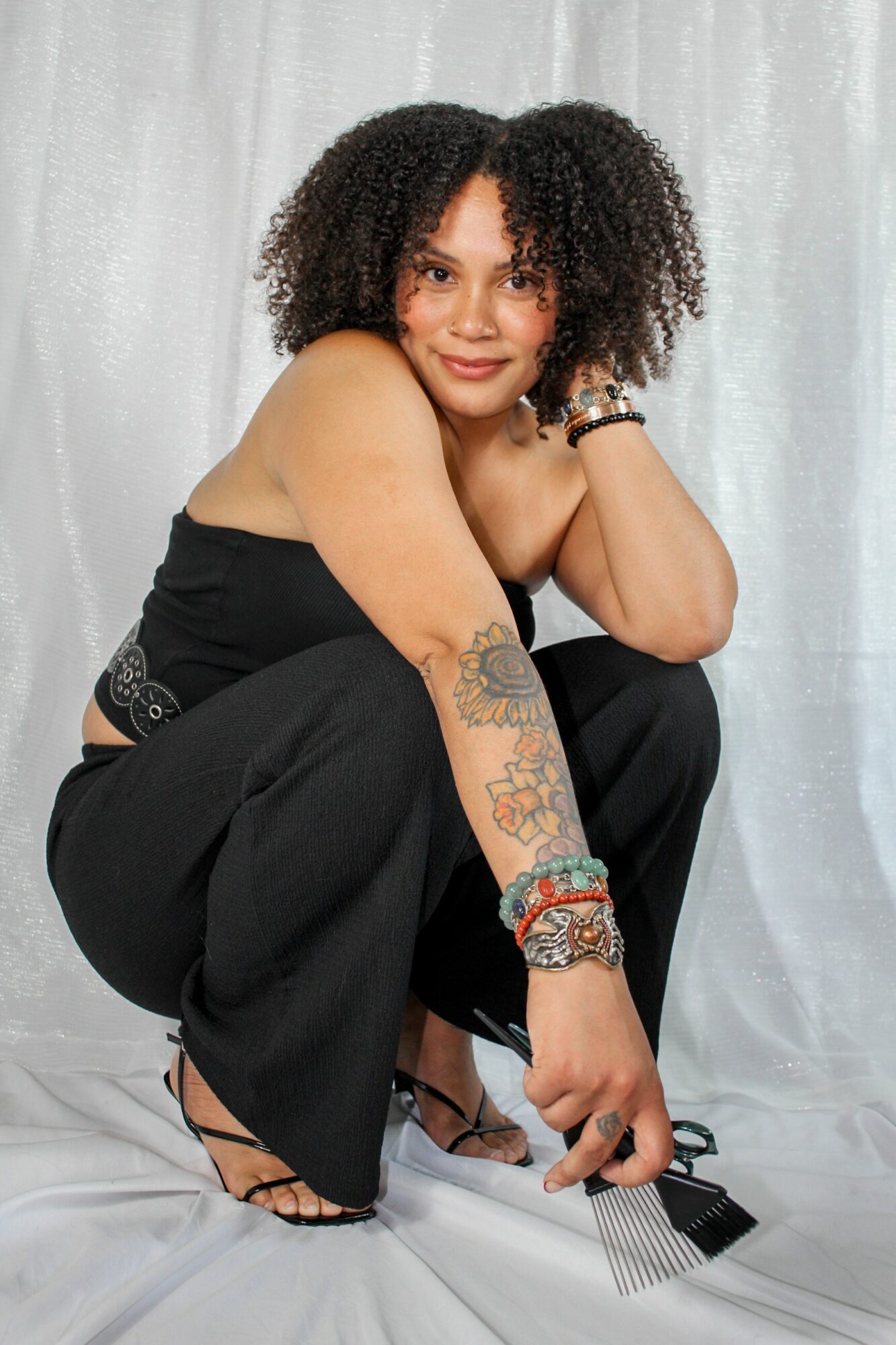 Woman with curly hair crouching, wearing a black top and pants, with tattoos and bracelets, against a light background.