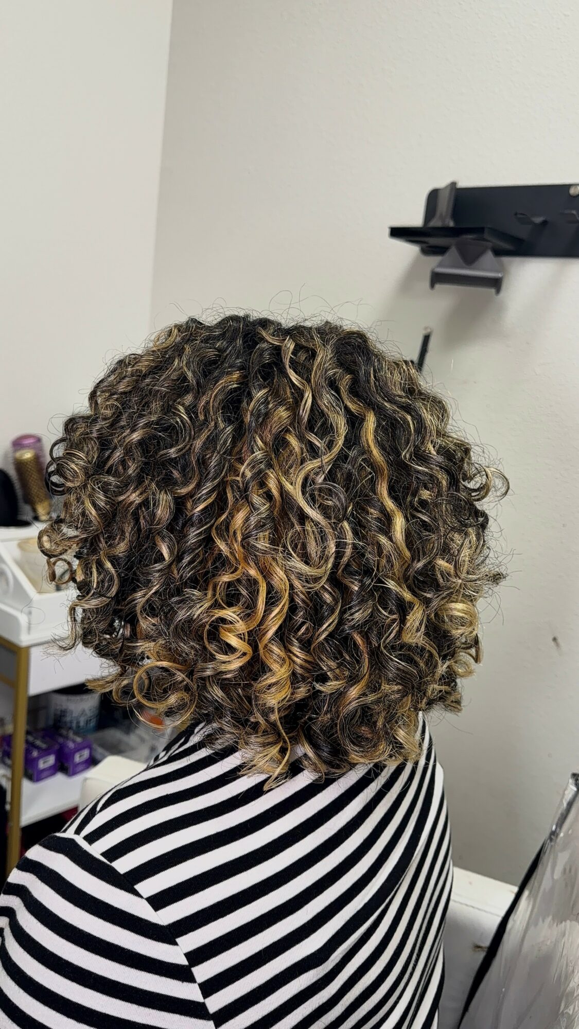 Back view of a person with curly, shoulder-length hair, wearing a black and white striped shirt, in a room with a white wall.
