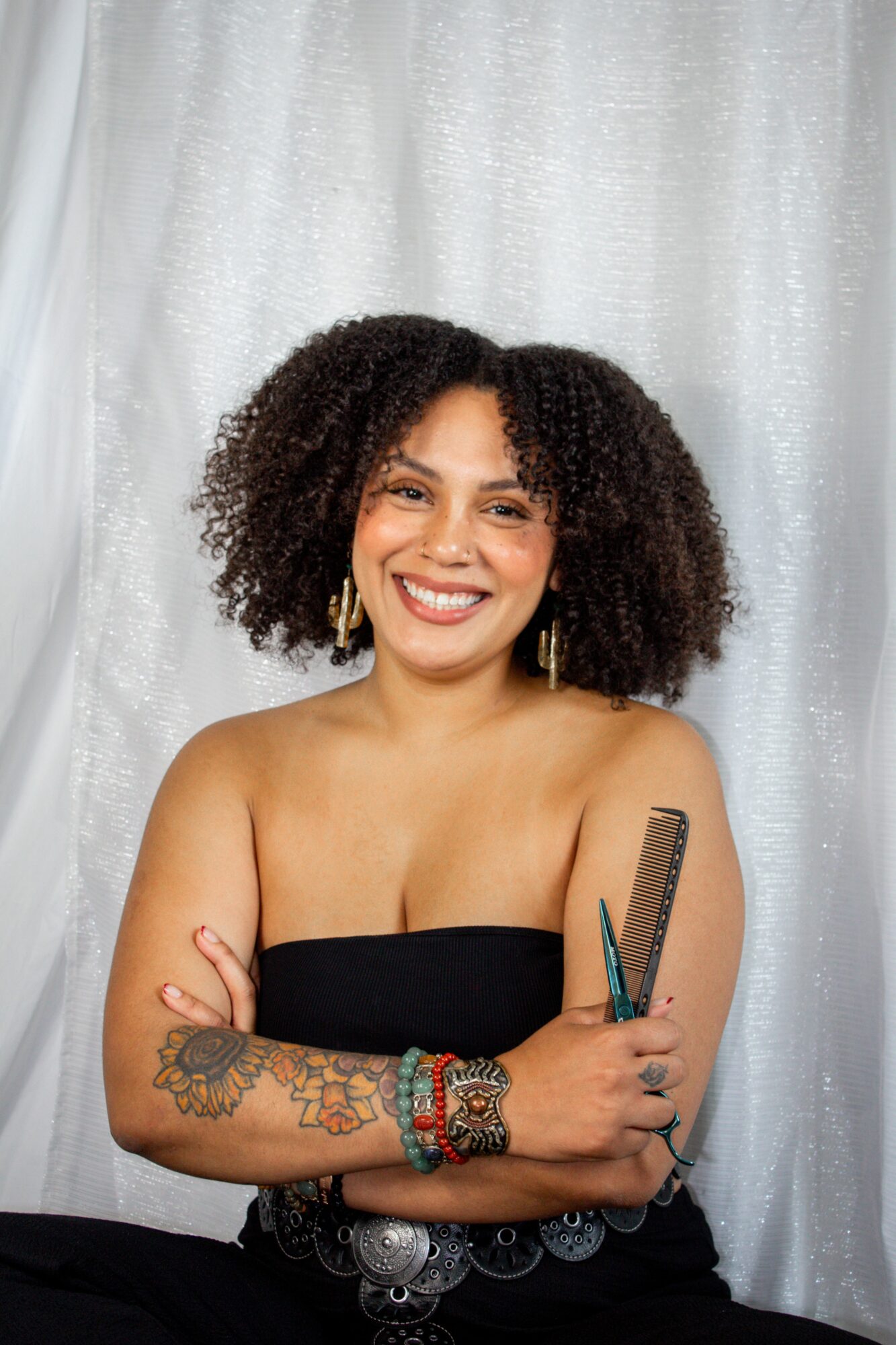Smiling woman with curly hair holding a comb and scissors, standing in front of a white curtain, arms crossed, wearing a strapless top.
