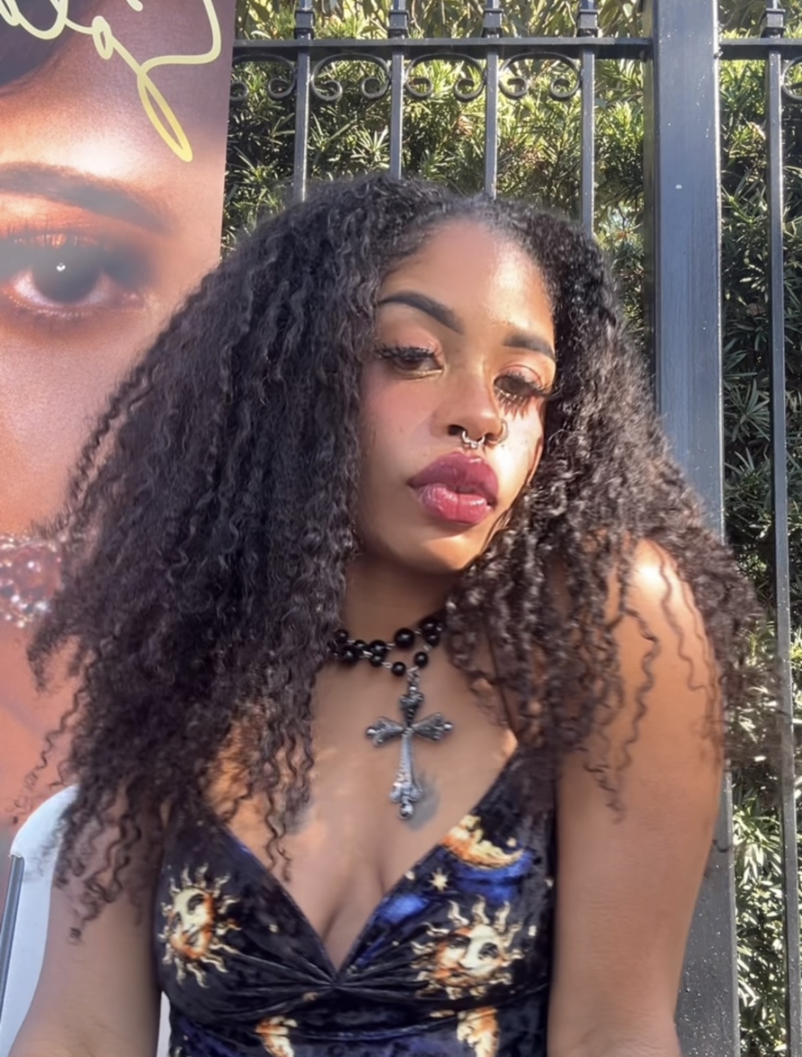 Young woman with curly hair, wearing a floral dress and jewelry, outdoors with a fence and greenery behind her.