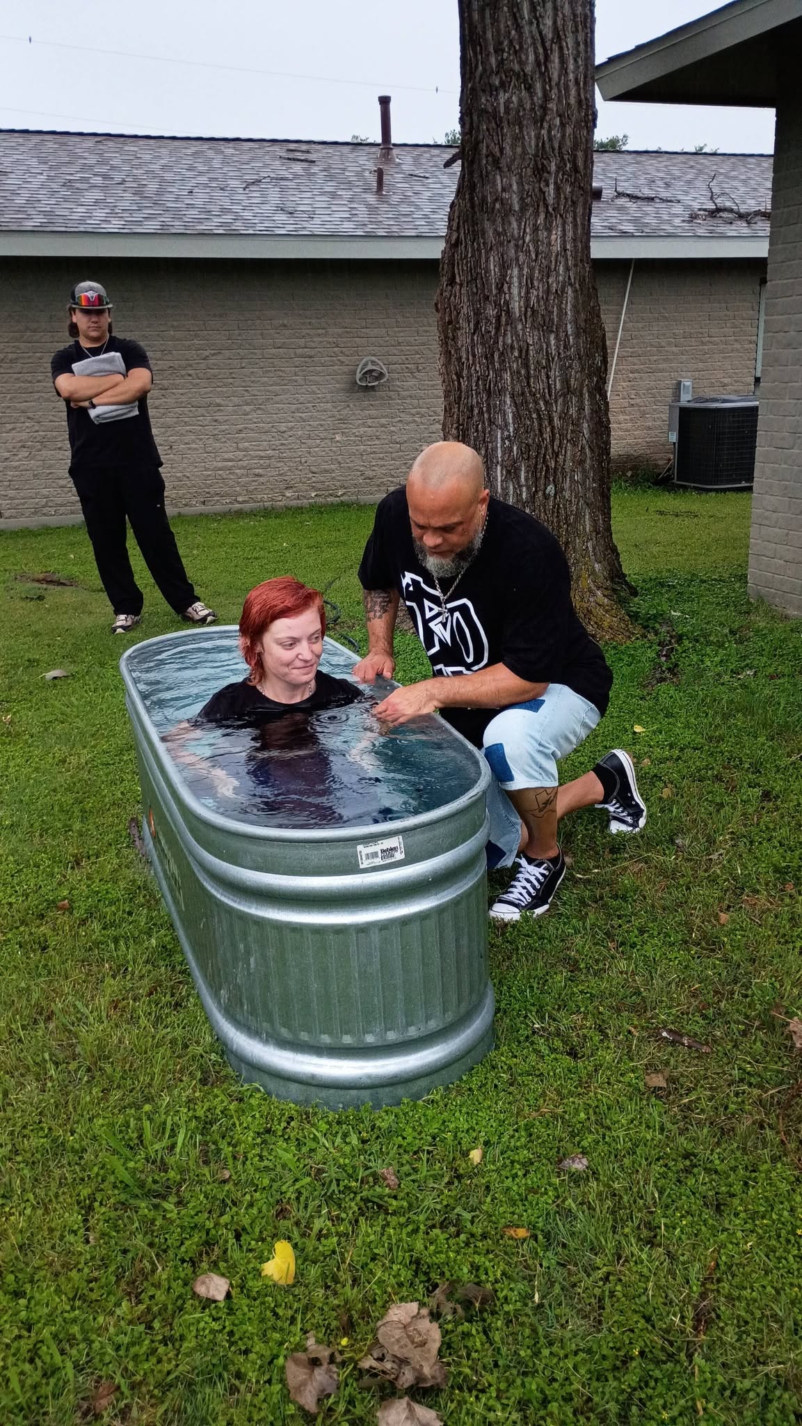 Person in a small pool outdoors with a man kneeling beside, another person standing nearby, grass and a building in background.