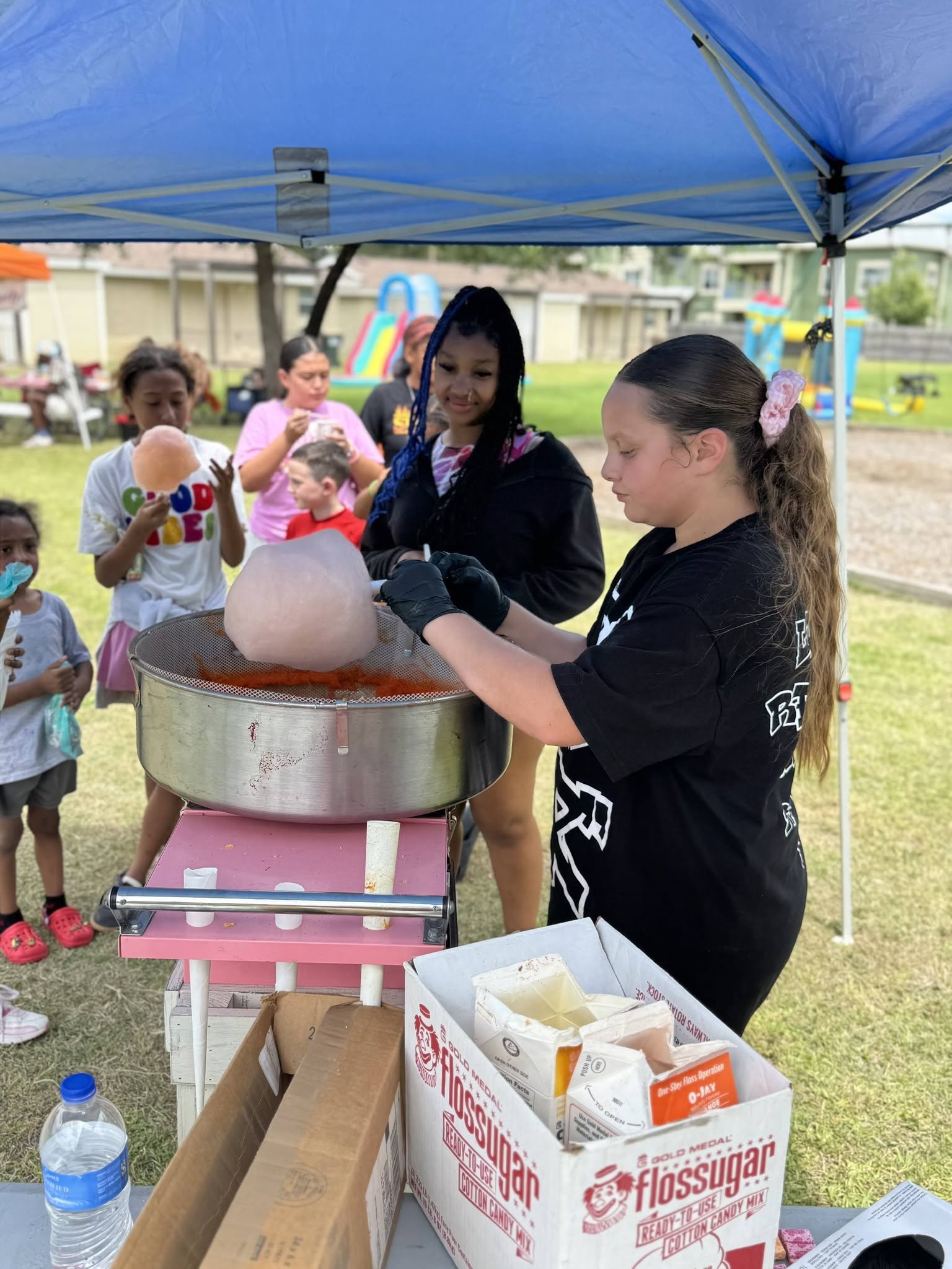 Two girls prepare cotton candy at a fair under a blue canopy, with children watching nearby outdoors.