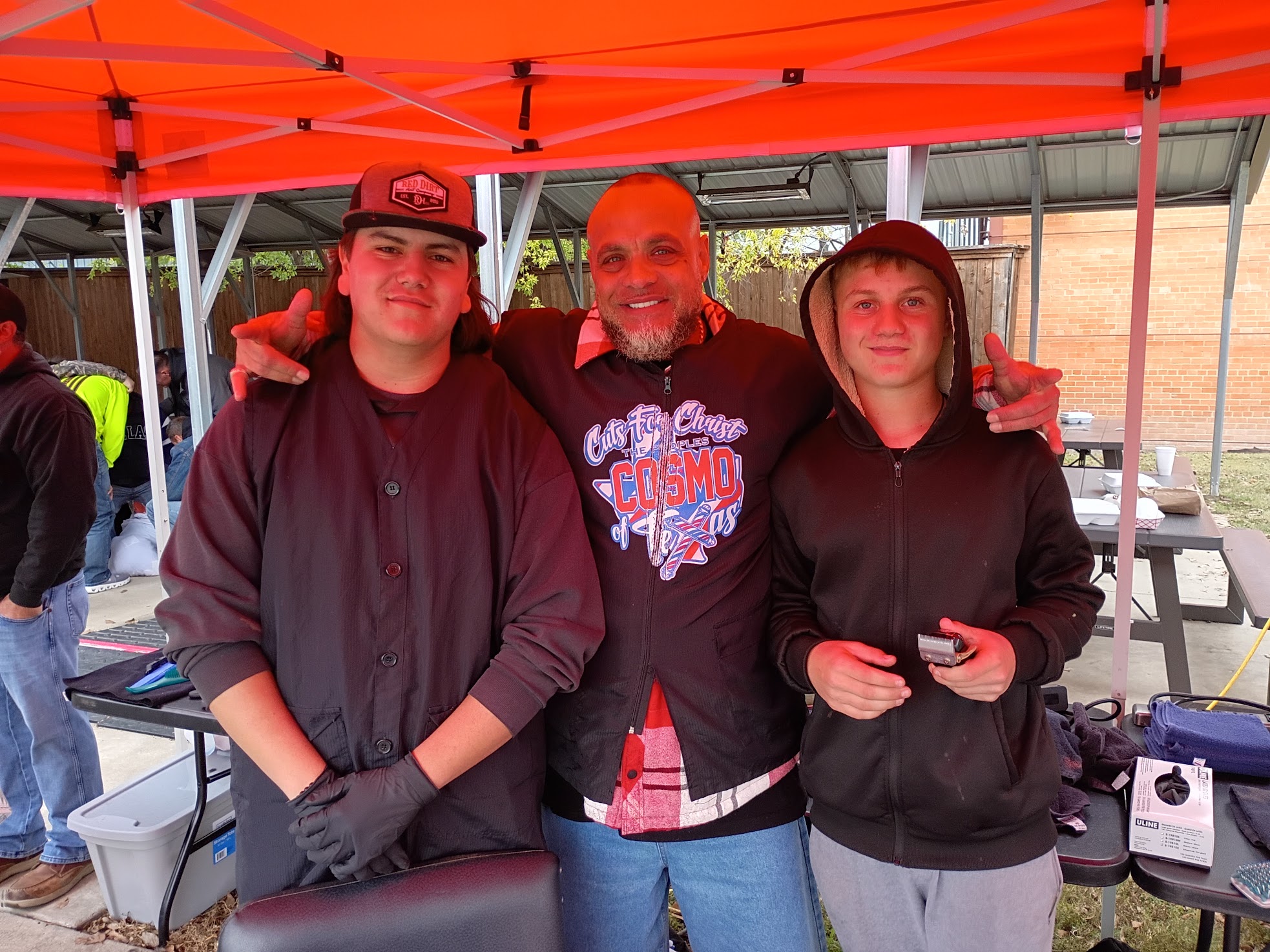 Three people standing under an orange canopy, smiling, with a table and equipment nearby.