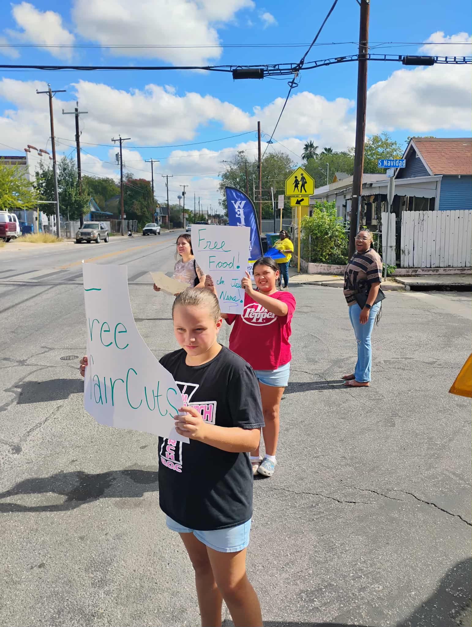 Group of children and a woman holding signs on sidewalk, with a street and blue sky in background.