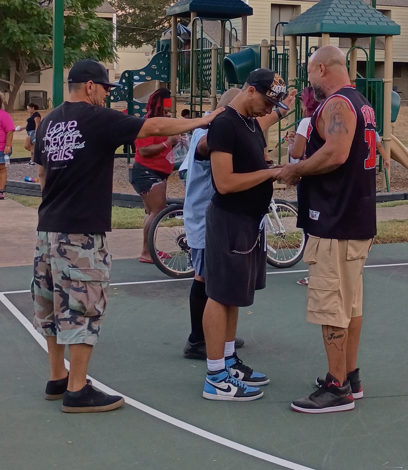 Three people standing on a playground court, two men and a woman, engaged in conversation, with playground equipment in background.
