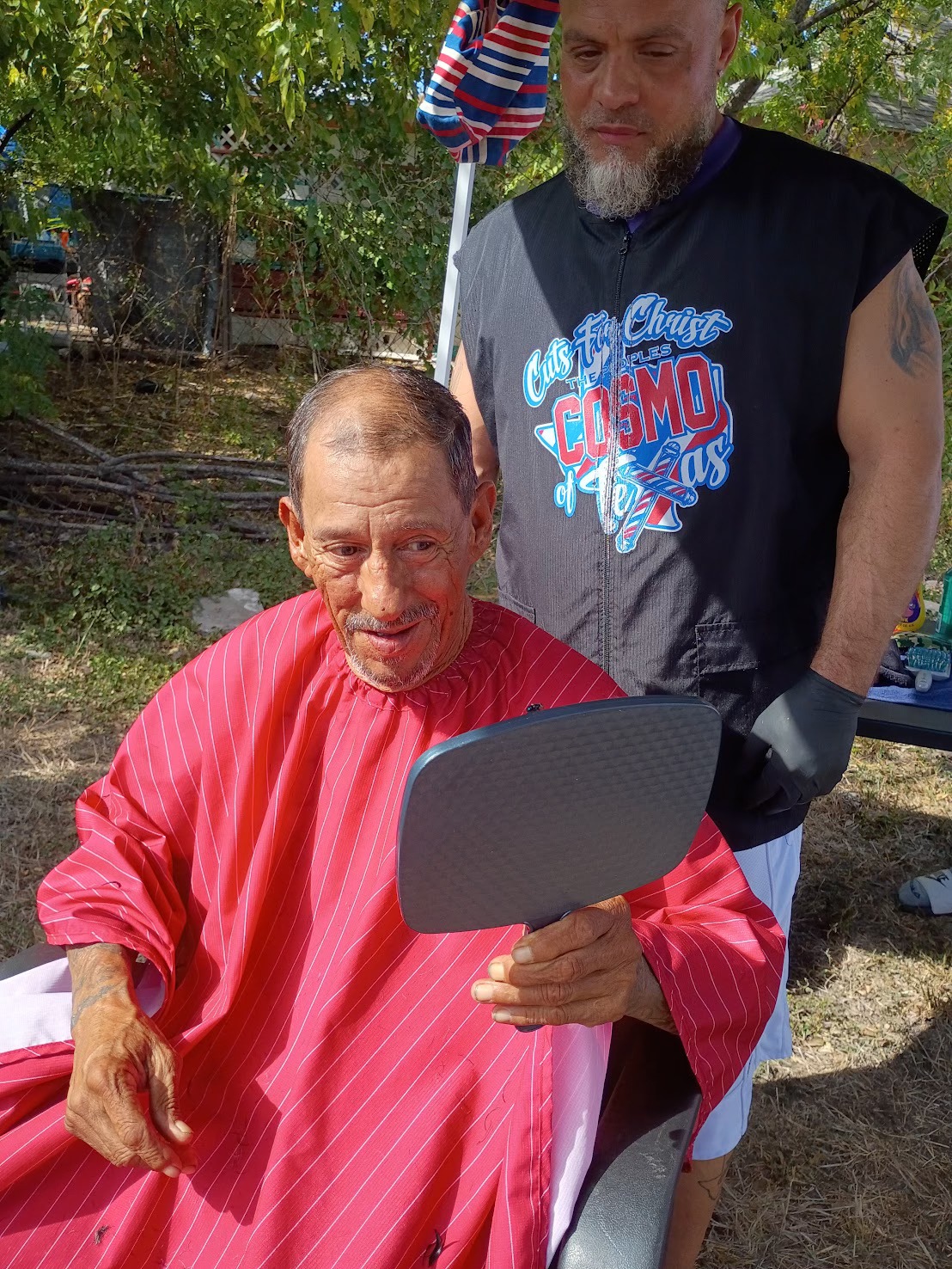 Two men outdoors, one seated with a red cape, the other standing behind, both looking at a mirror.