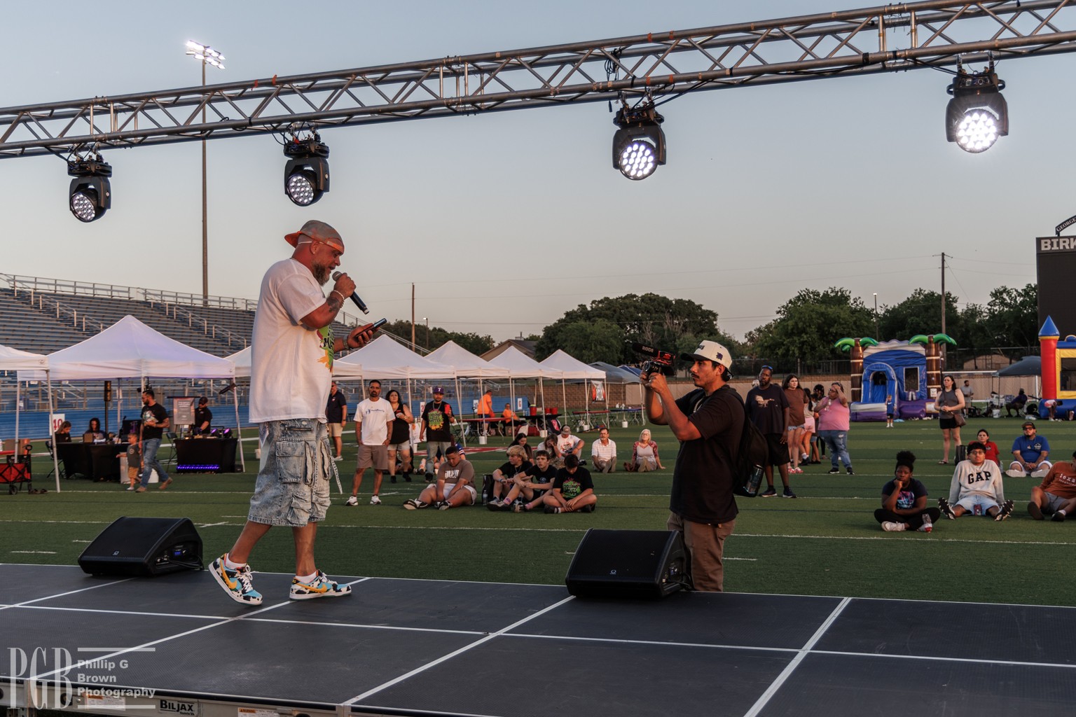 A man in a white shirt and shorts speaks into a microphone on a stage, with a person in black nearby, at an outdoor event.