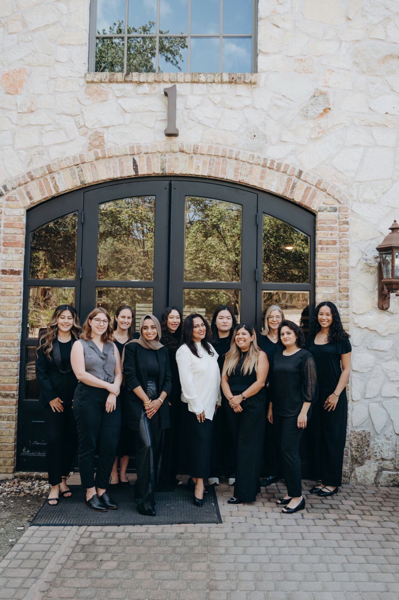 Group of women standing in front of a large arched glass door, stone wall background, outdoors.