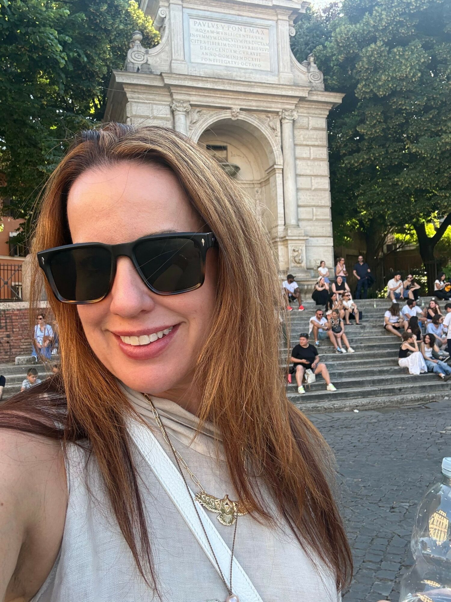 Woman with sunglasses smiling, standing outdoors near a stone building with stairs and people in the background.