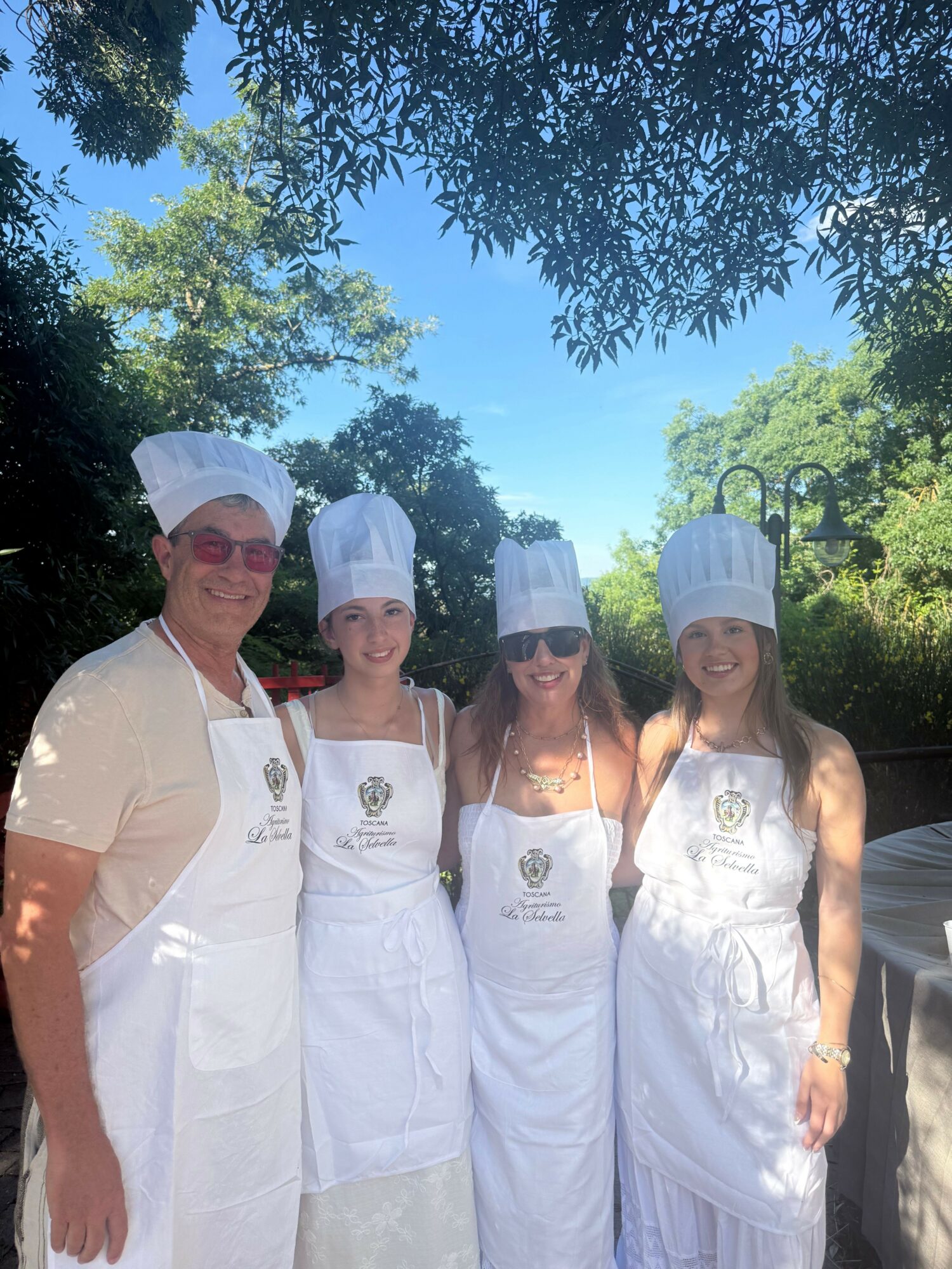 Four women in white dresses and chef hats standing outdoors with trees and blue sky in background.