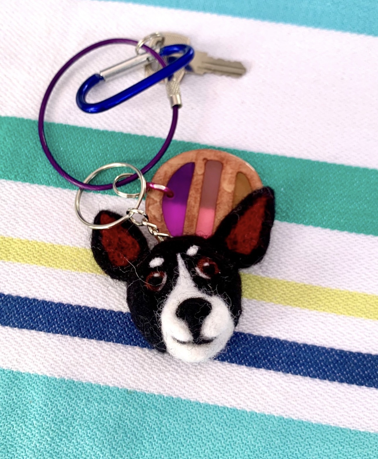 Keychain with a dog's face, a pink and orange round charm, and a blue paperclip, on a striped fabric background.