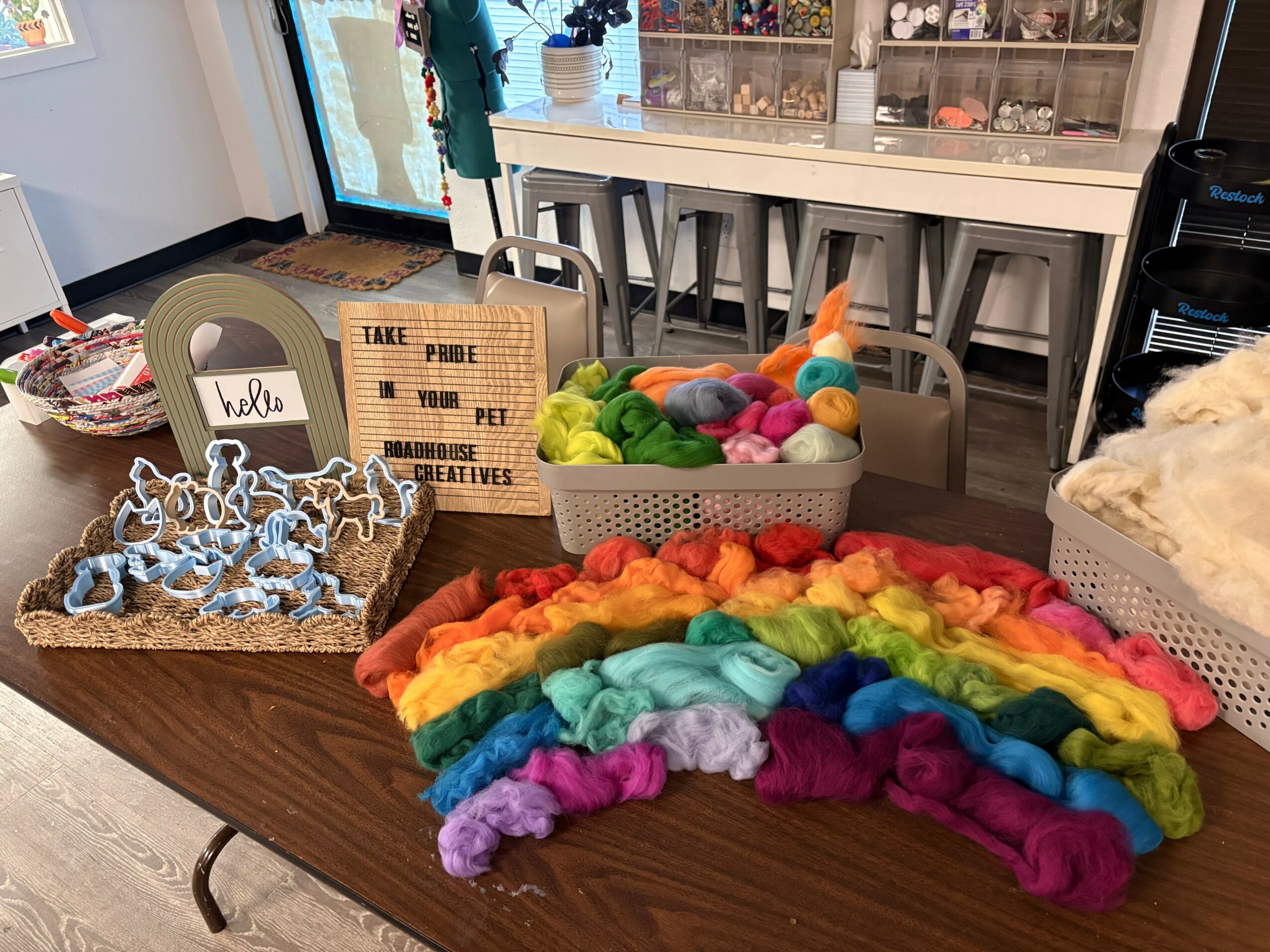 Table with colorful yarn and felted wool, background shows a cozy room with a sign and shelves.