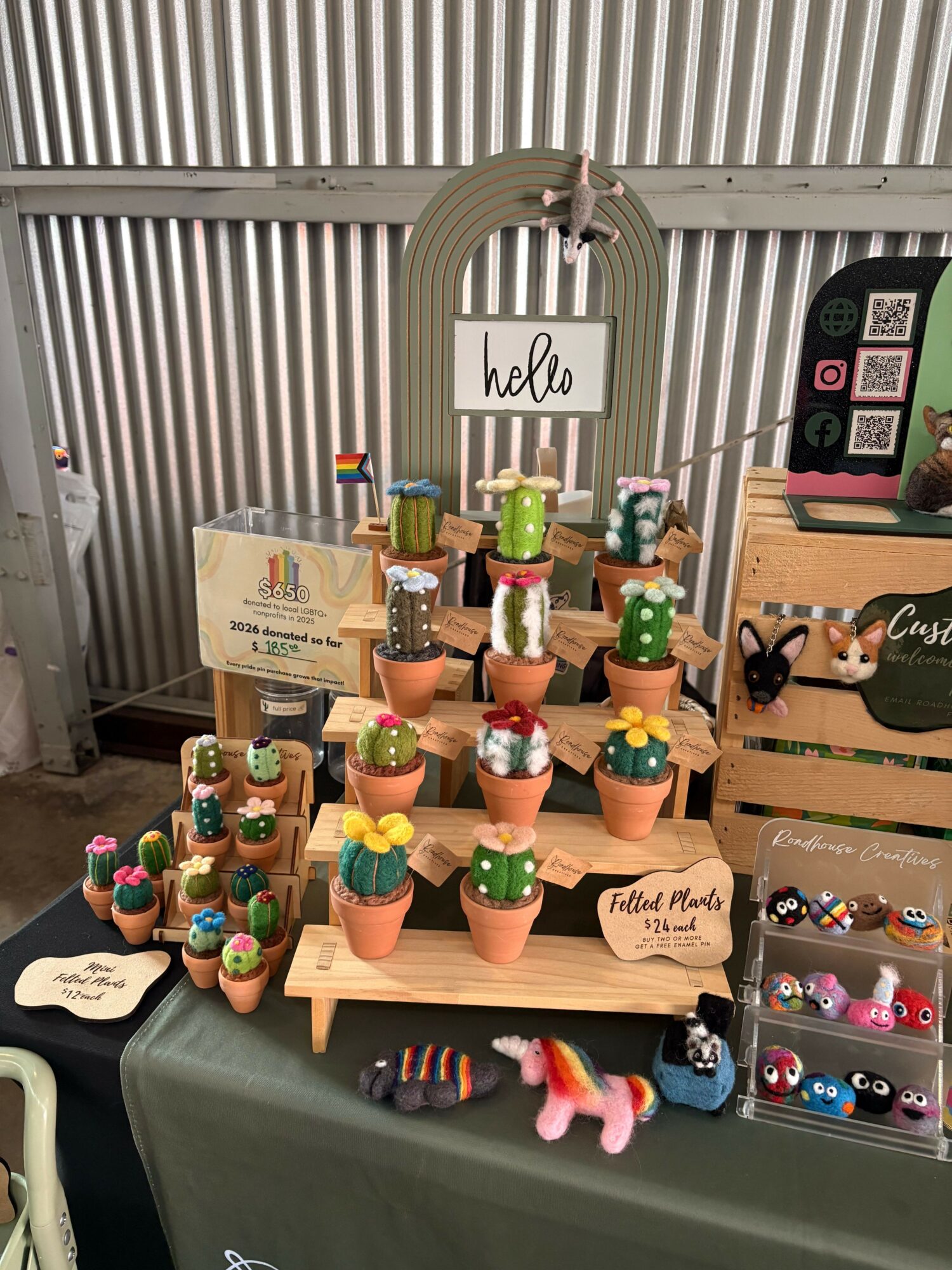 Display of colorful handcrafted figurines on a table with a sign and a green tablecloth, set against a corrugated metal background.