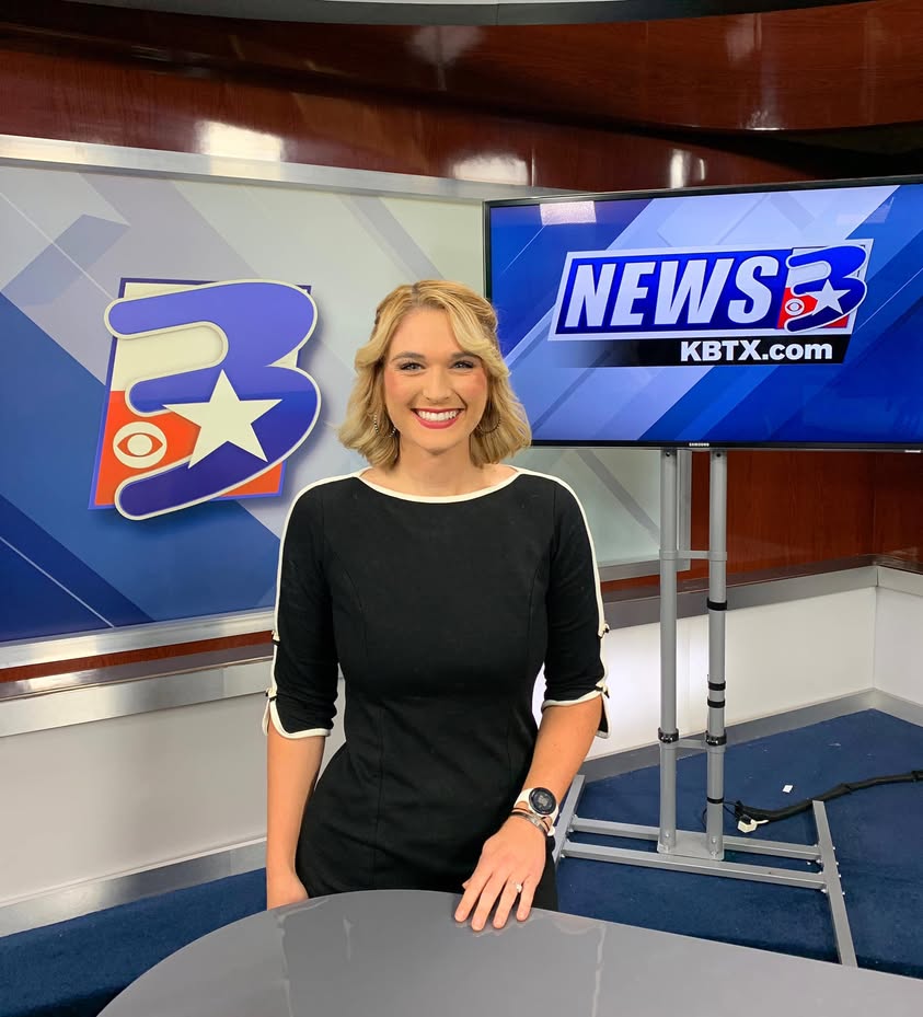 Woman with blonde hair smiling in a news studio, standing in front of screens displaying news logos.