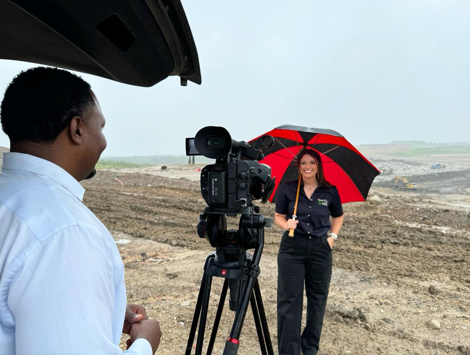 Woman holding a red and black umbrella being filmed outdoors with a camera and a man in the foreground.