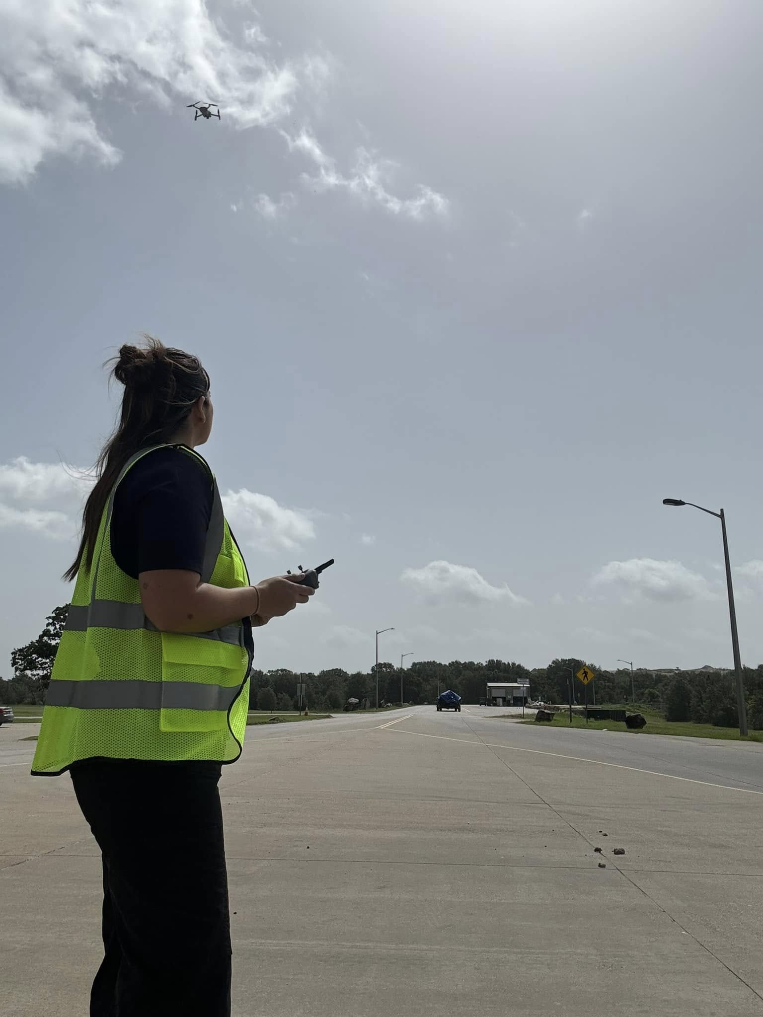 Person wearing a yellow safety vest standing on a road, looking at a drone in the sky, holding a device.