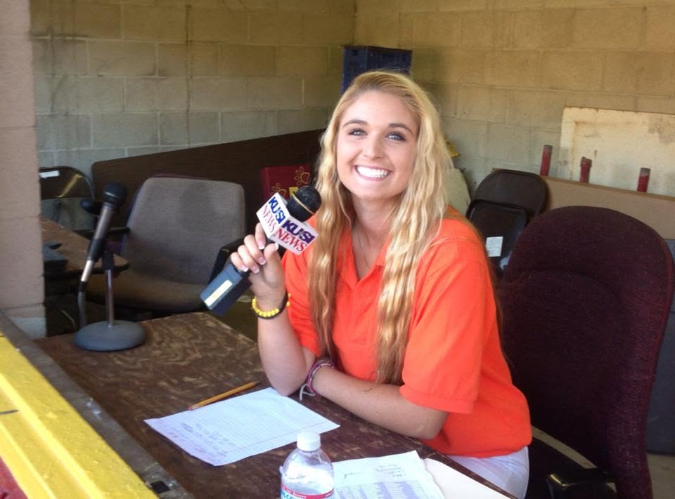 Young woman with long blonde hair smiling, holding a microphone, sitting at a table with papers and a water bottle.