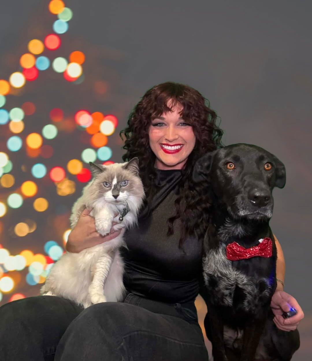 Woman smiling with a cat and a dog, Christmas tree with colorful lights in background.