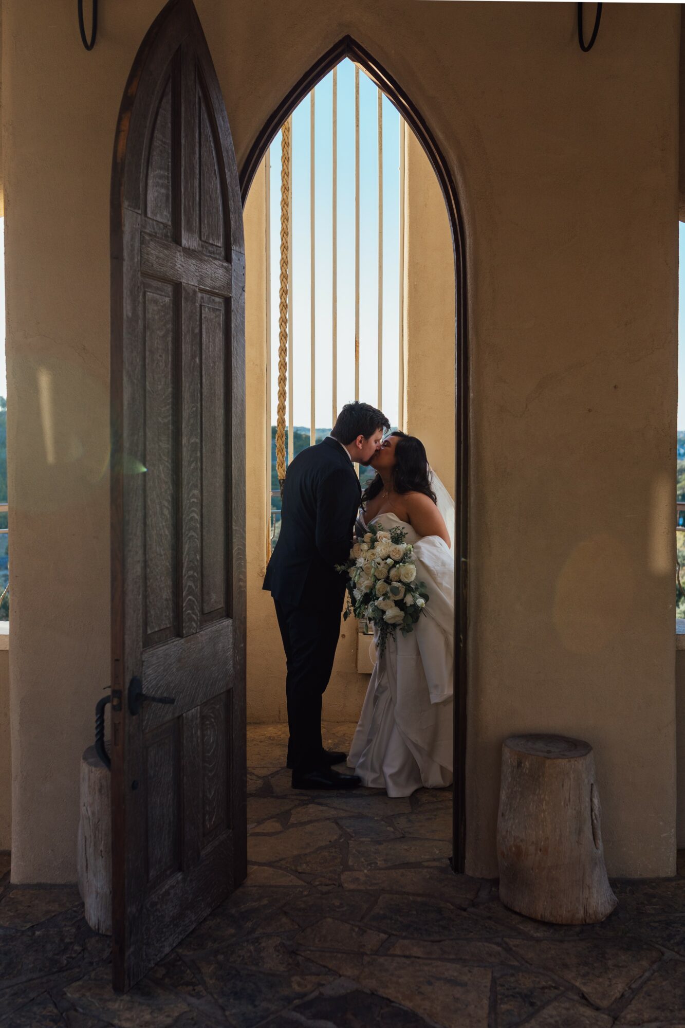 A couple stands close together in a doorway, with a window and bars behind them, holding a bouquet of flowers.