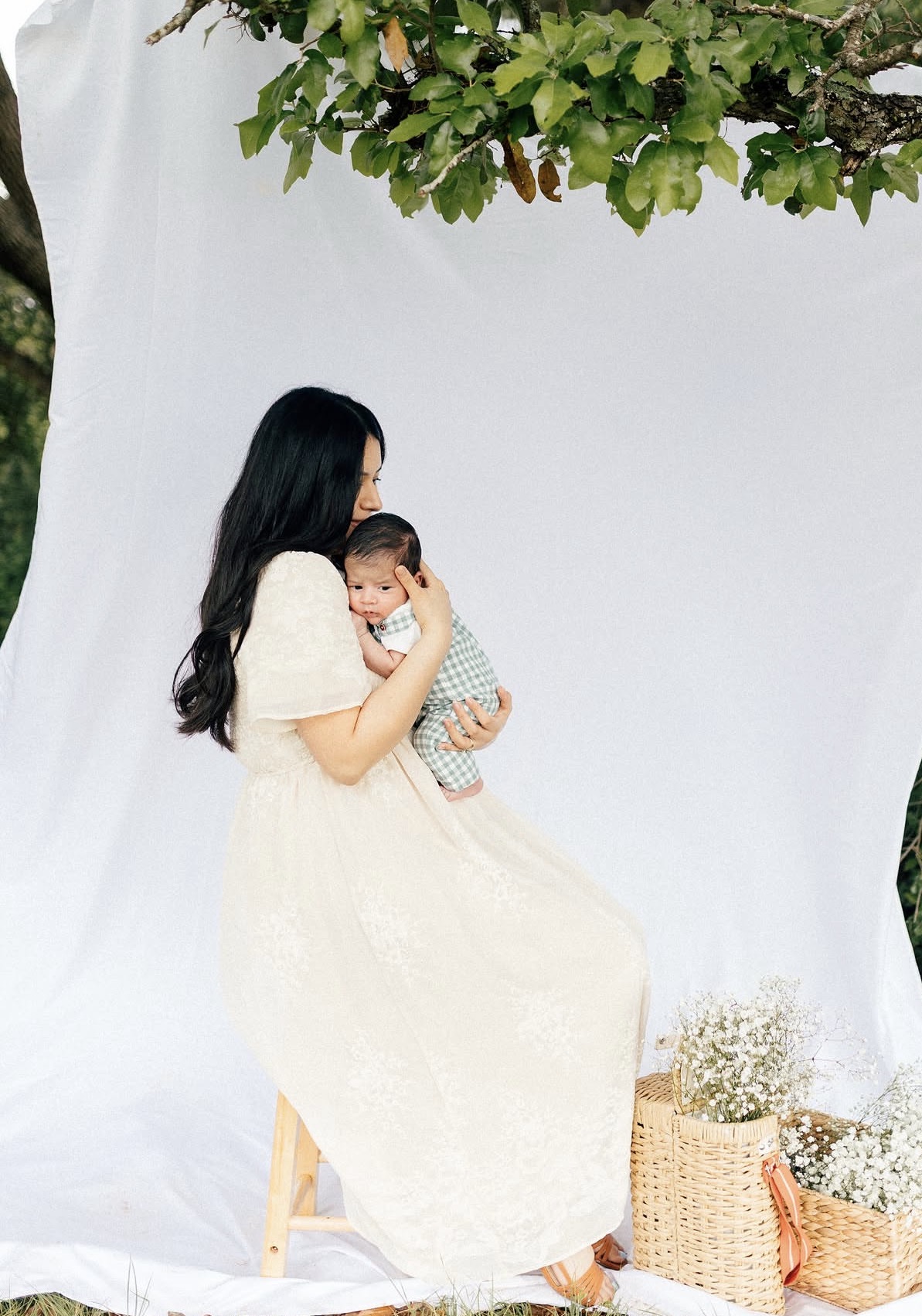 Woman in a light-colored dress holding a baby, sitting on a stool outdoors with a white backdrop and greenery.