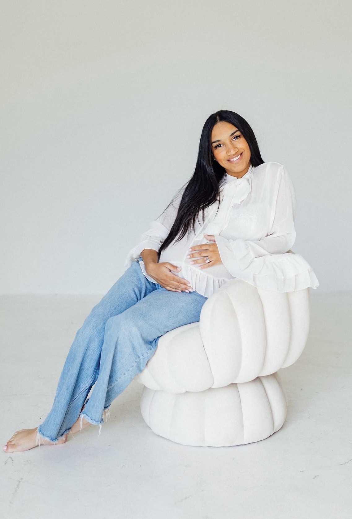 Woman with long dark hair sitting on a white sculptural chair, smiling, wearing a white blouse and blue jeans, against a plain background.
