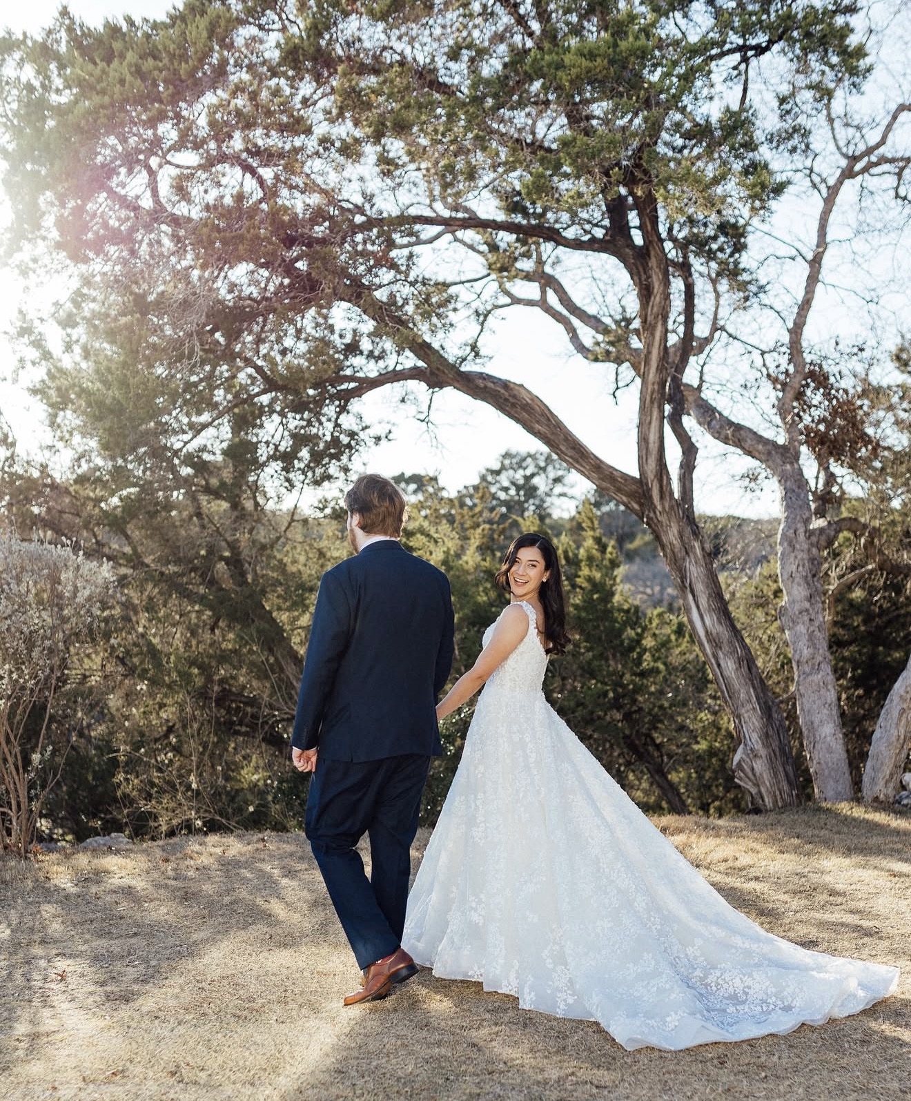 A bride and groom stand outdoors under a large tree, with the bride in a white wedding dress and the groom in a suit.