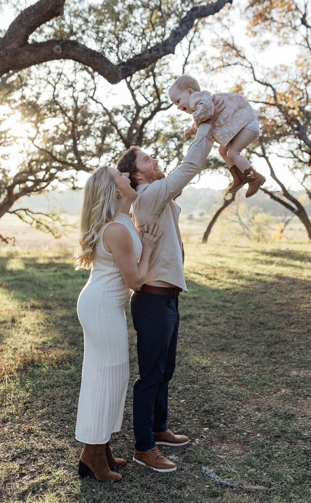 A family outdoors with a woman lifting a child in the air, trees and sunlight in the background.