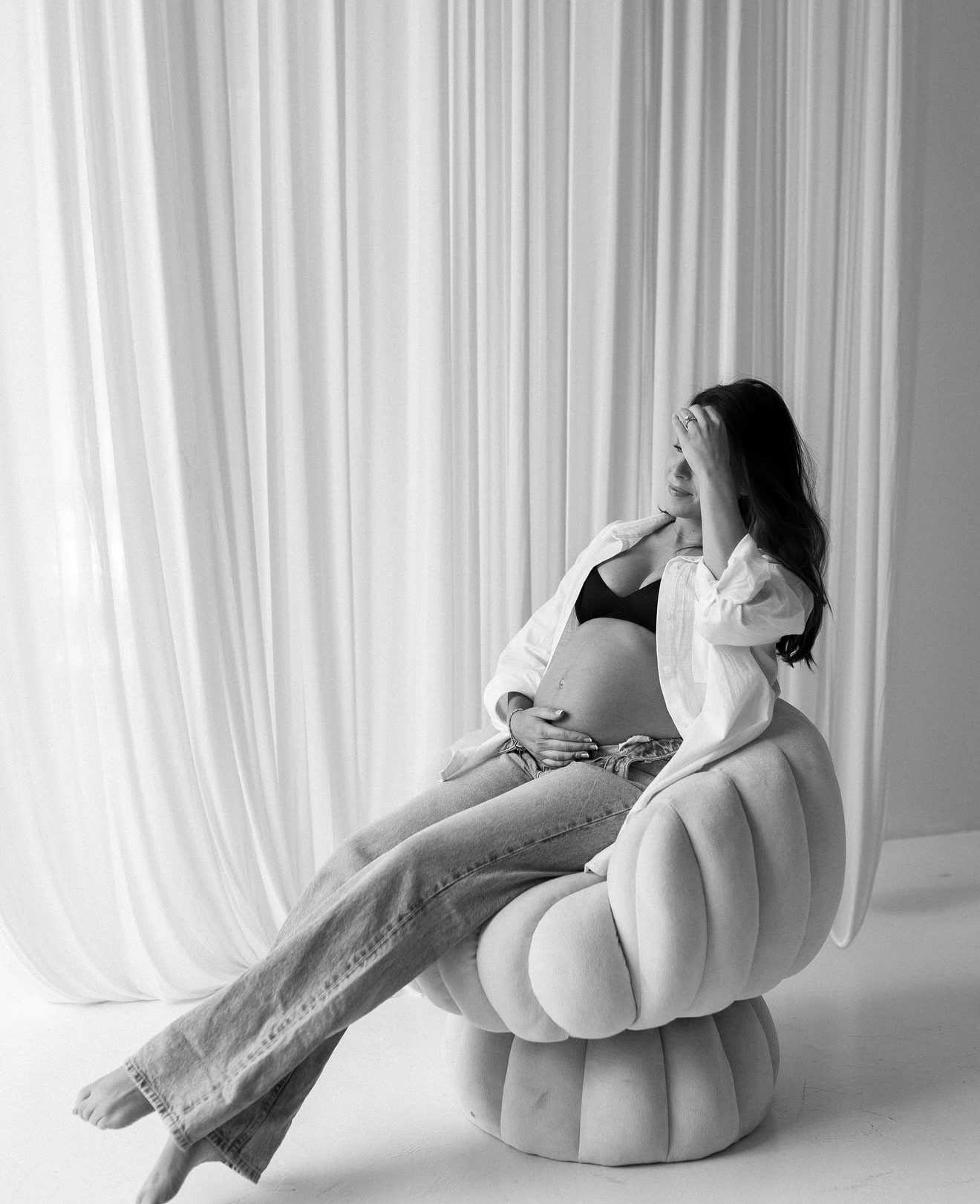 Pregnant woman sitting on a rounded chair, covering her face with her hand, in front of curtains, in black and white.