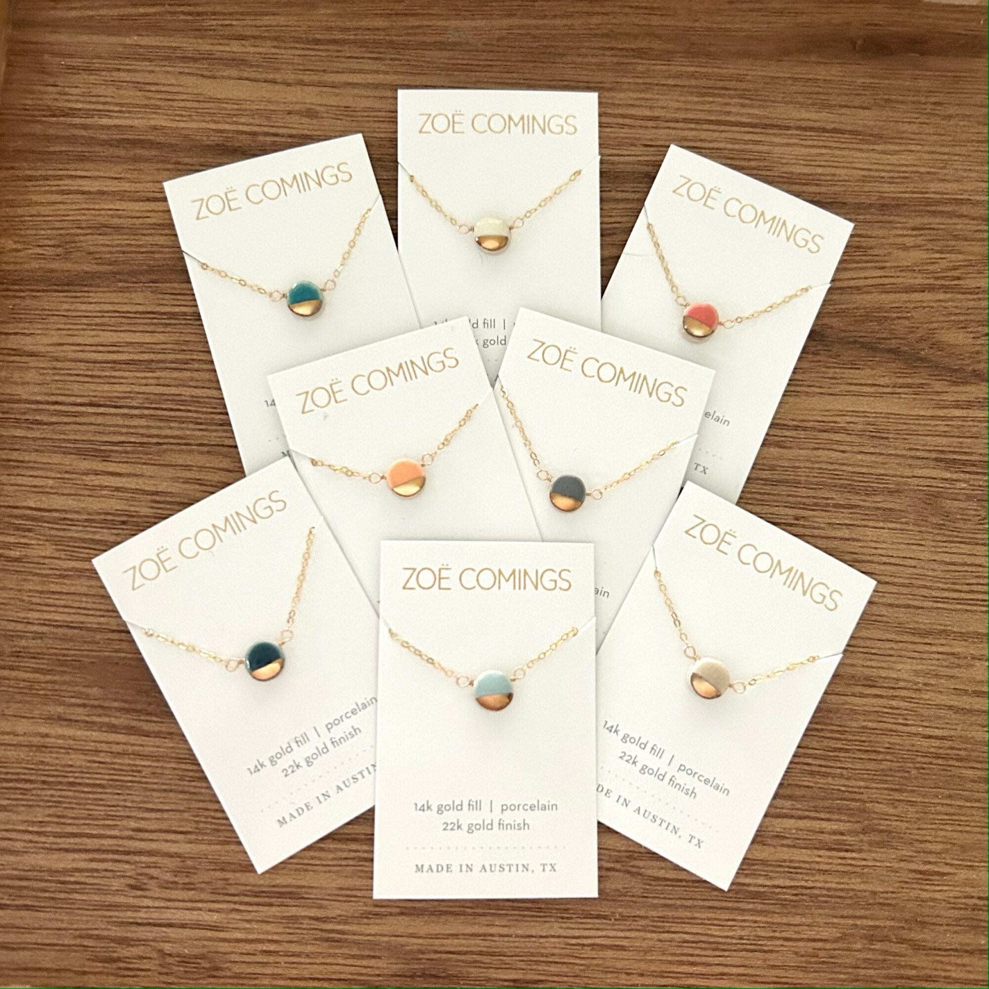 Seven necklaces with colorful pendants on white cards, arranged on a wooden surface. Each card has the brand name 'ZOE COMINGS'.