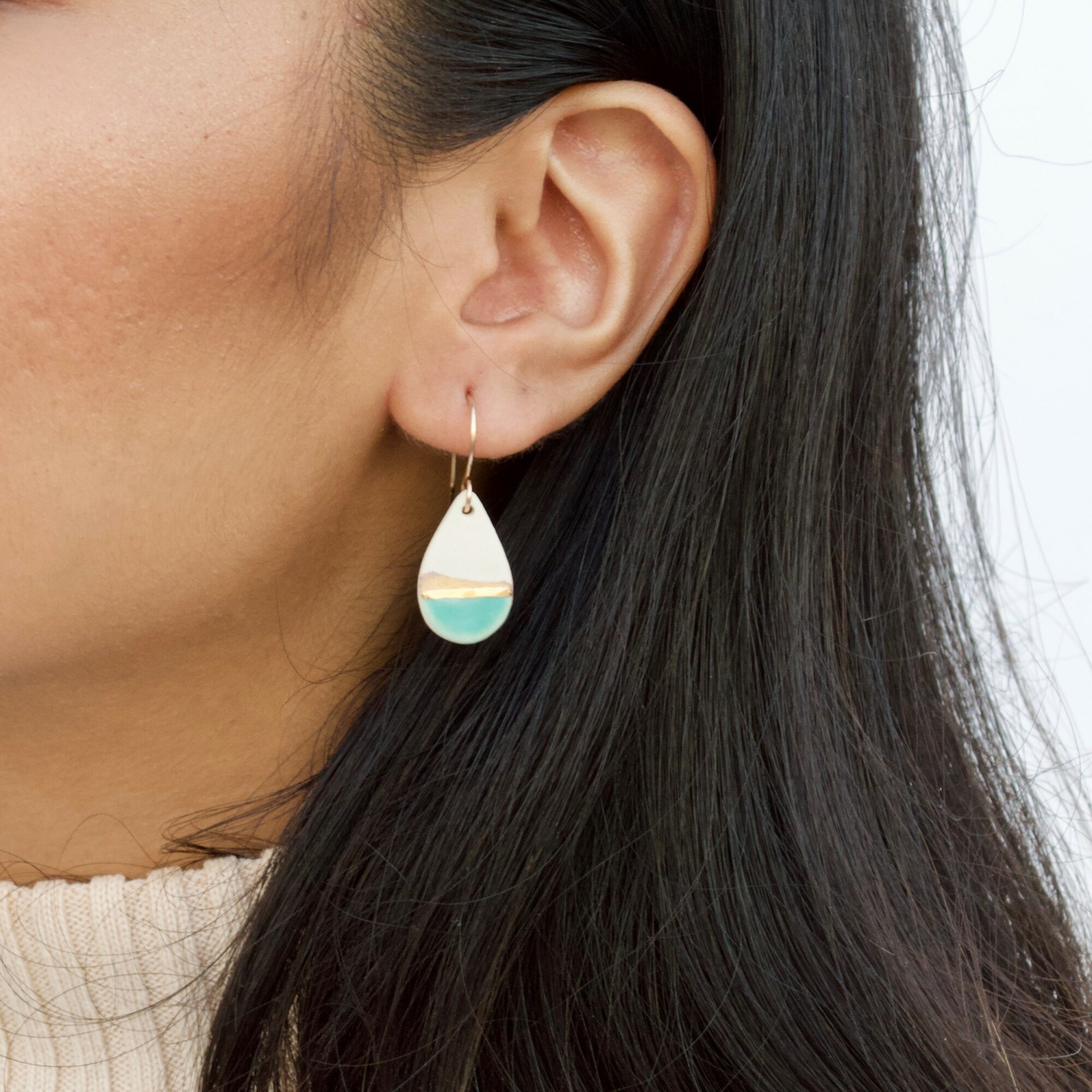 Close-up of a woman's ear with a teardrop-shaped earring featuring a blue and white design, black hair, and a beige top.