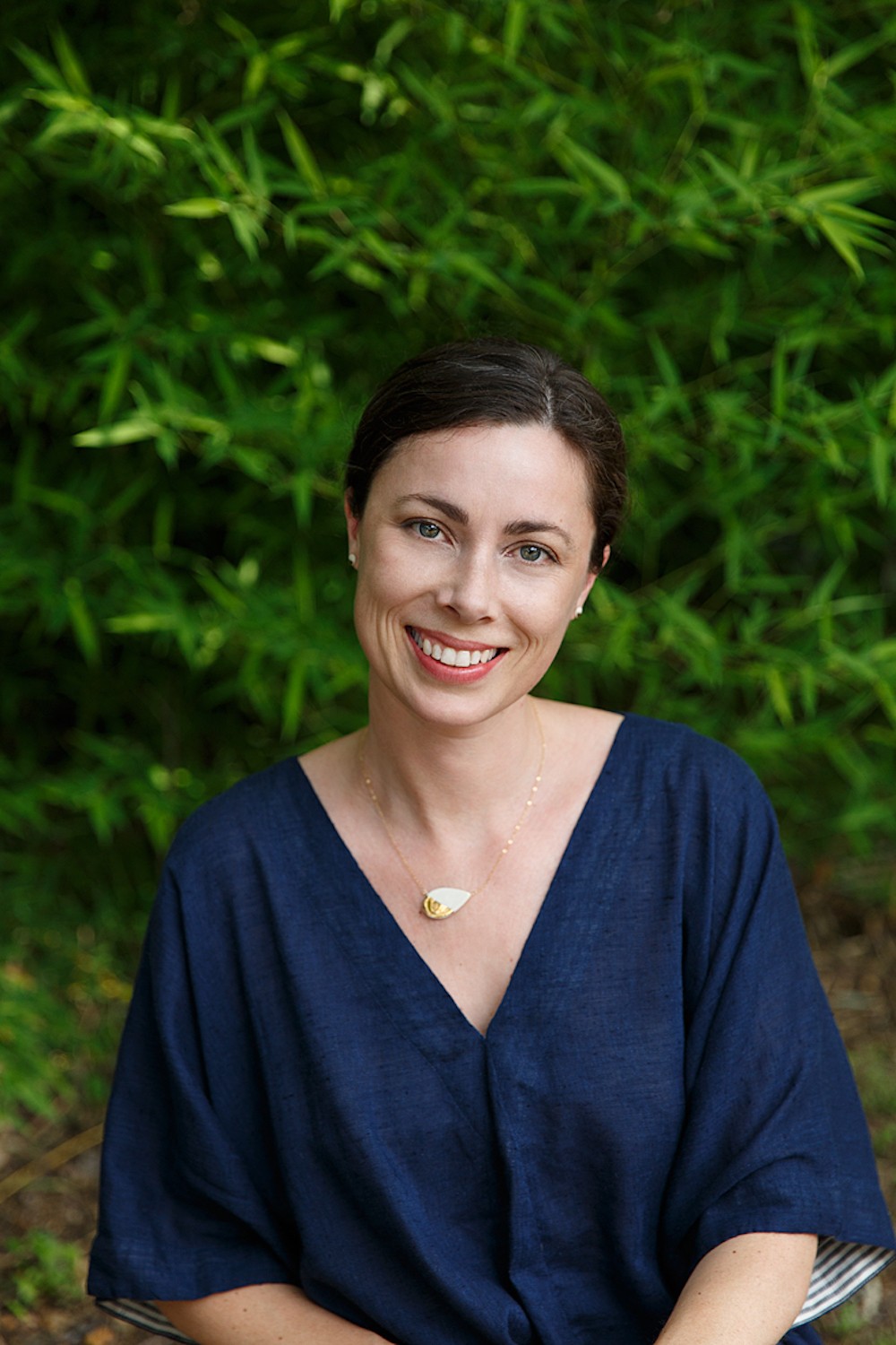Smiling woman with short dark hair wearing a navy blue top and a necklace, standing outdoors with green foliage background.