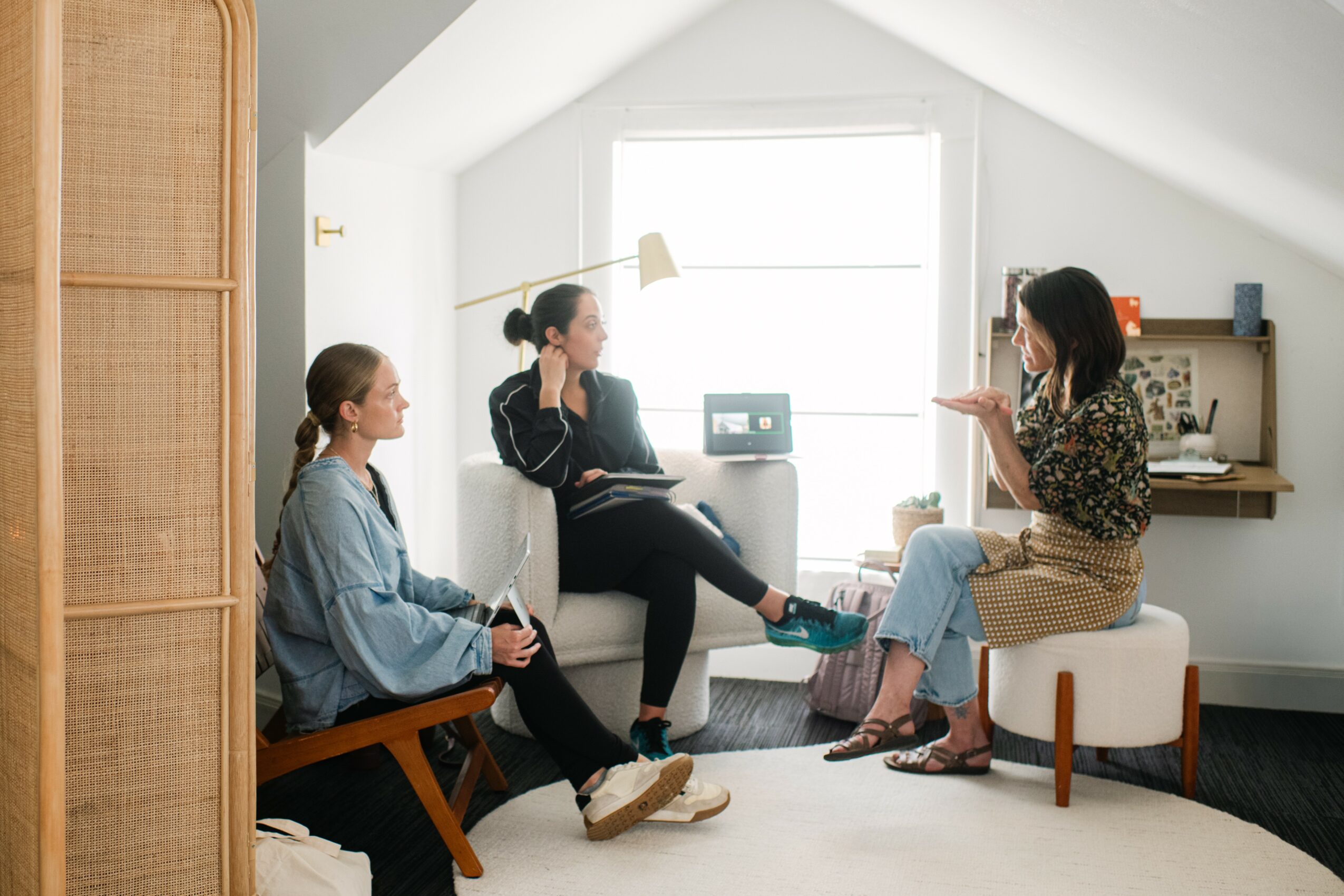 Three women sitting in a bright room having a conversation, with a window and shelves in the background.