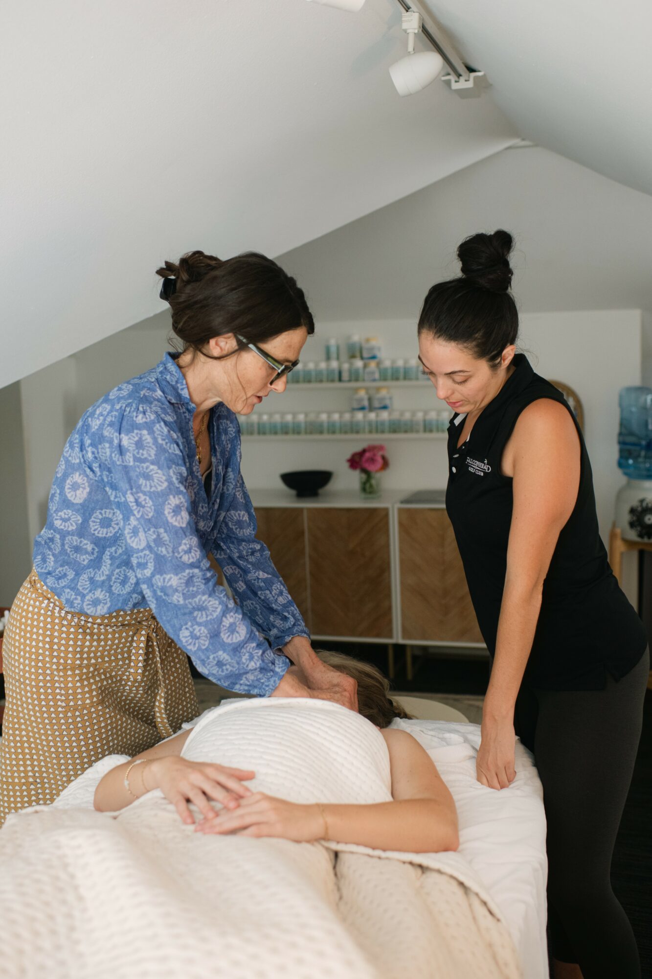 Two women assist a person lying in bed, one adjusting the blanket, in a room with shelves and a water cooler.