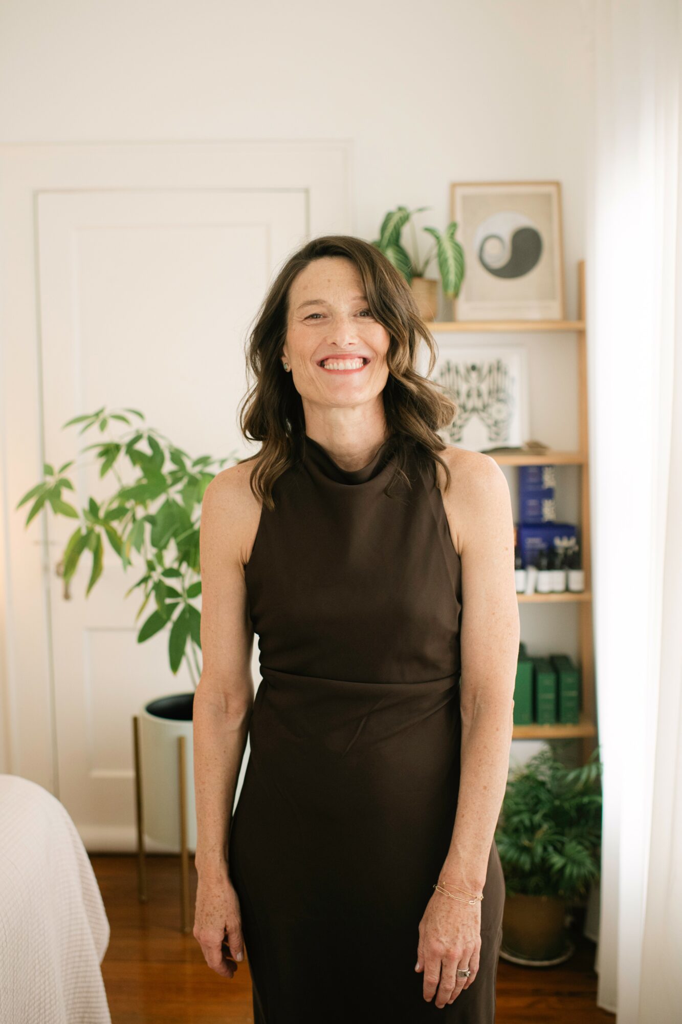 Woman with shoulder-length dark hair smiling, wearing a sleeveless black dress, standing indoors with plants and shelves in background.