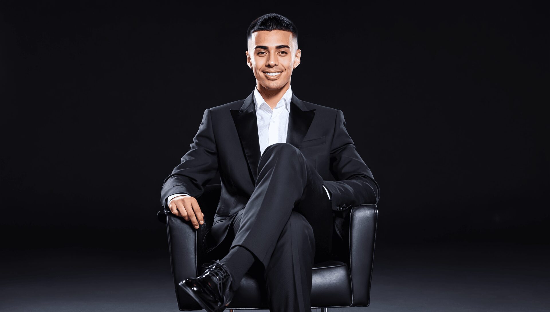 Young man in a suit sitting on a black chair, smiling, against a dark background.