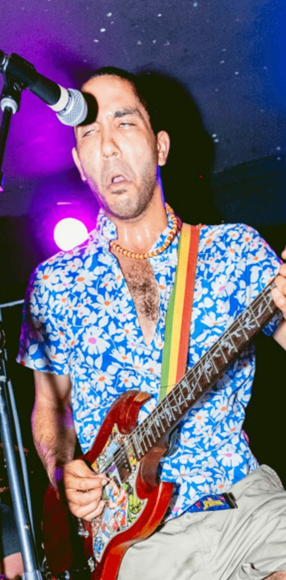 Person with long hair playing an electric guitar, wearing a blue floral shirt, in a performance setting.