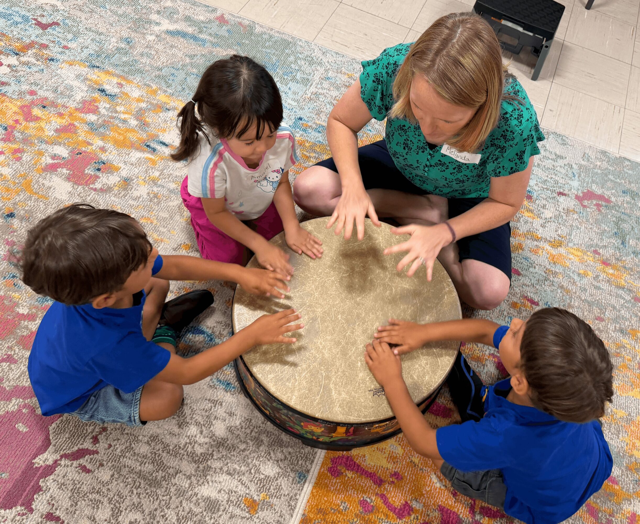 Adult and three children sit around a round drum, playing with their hands on it, on a colorful rug.