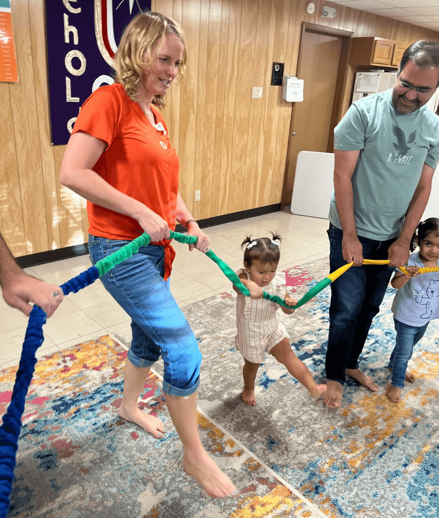 Children and adults hold a colorful rope indoors, participating in a group activity, with a wooden wall and a banner in the background.
