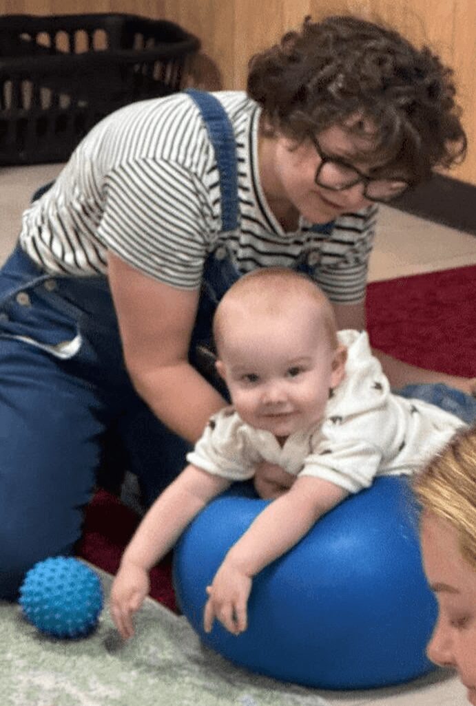 Person with curly hair and glasses leaning over a baby lying on a blue exercise ball, smiling, with a textured blue ball nearby.