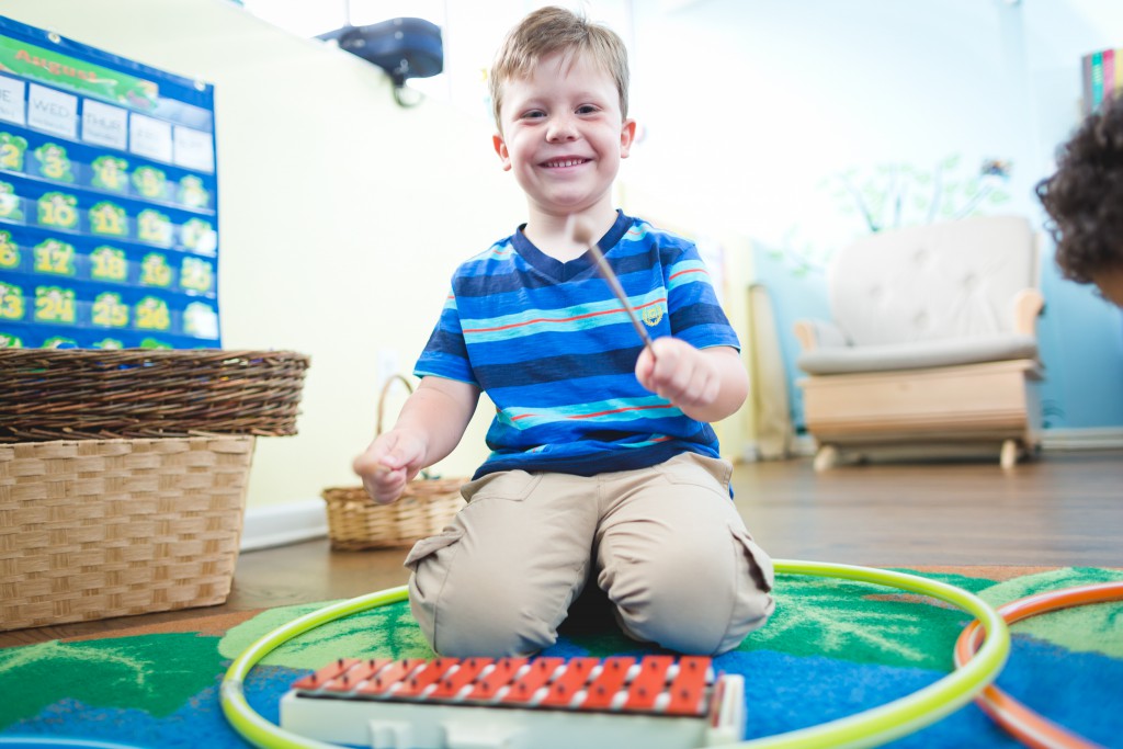 Child sitting on a colorful mat, smiling, holding a stick, with a xylophone and hula hoop nearby, in a bright room.